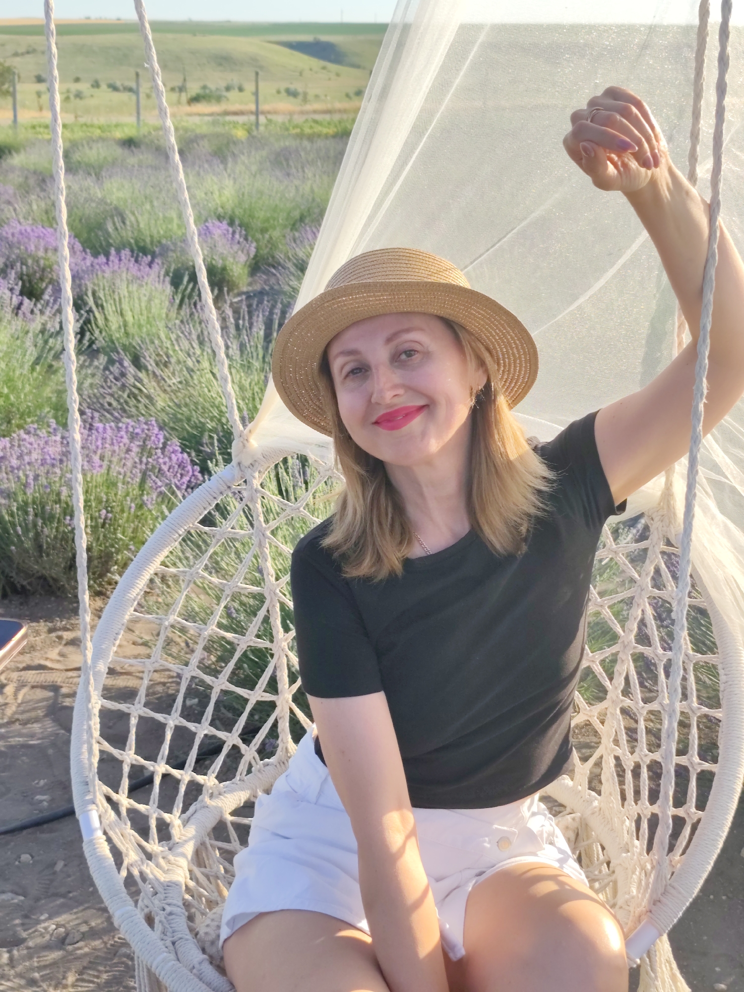 A woman with blonde hair and bright pink lipstick, wearing a straw hat and a black t-shirt, is sitting in a white macrame hanging chair. She is in a lavender field, with rows of purple lavender stretching into the background, under a clear sky. The sun is shining, casting a warm glow on the scene, suggesting it is a sunny day, likely during golden hour due to the soft lighting. The woman is smiling and looking towards the camera, with one hand raised, showing a ring on her finger. The setting appears to be a tranquil, scenic outdoor location, possibly a lavender farm, given the abundance of blooming lavender. The overall mood is relaxed and pleasant. No visible text is present in the image.