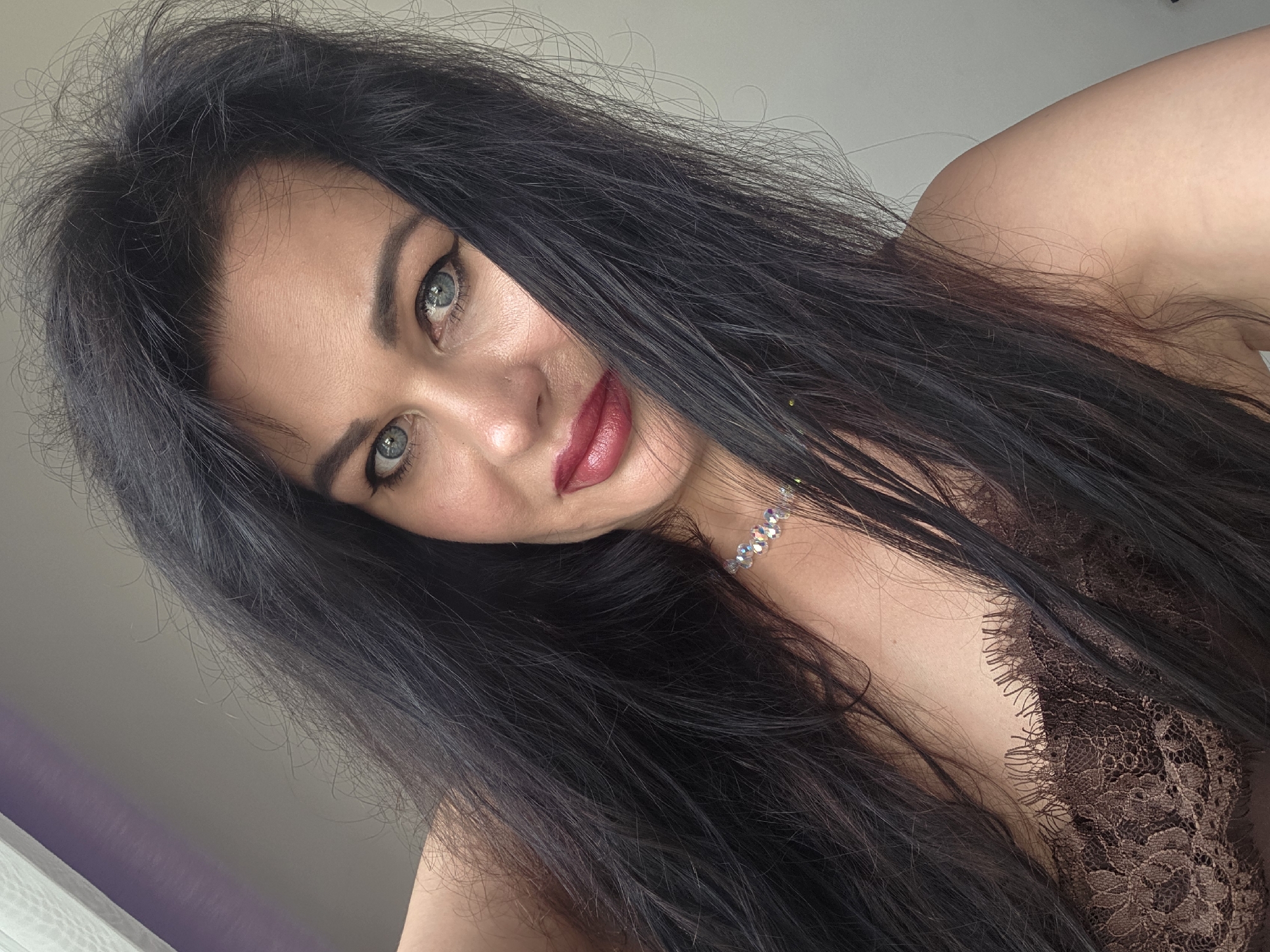 A close-up, slightly angled portrait features an adult female with long, dark, voluminous hair, which partially frames her face and shoulders. Her blue eyes, enhanced with dark eyeliner, look directly towards the camera. She wears dark red lipstick on full lips, exhibiting a subtle smile. A delicate, iridescent beaded choker necklace is visible on her neck, alongside a dark brown, floral lace garment. The background consists of a plain, light-colored wall, with a hint of a purple-tinted surface and a light-colored curtain or blind visible in the upper left corner, indicating an indoor location.