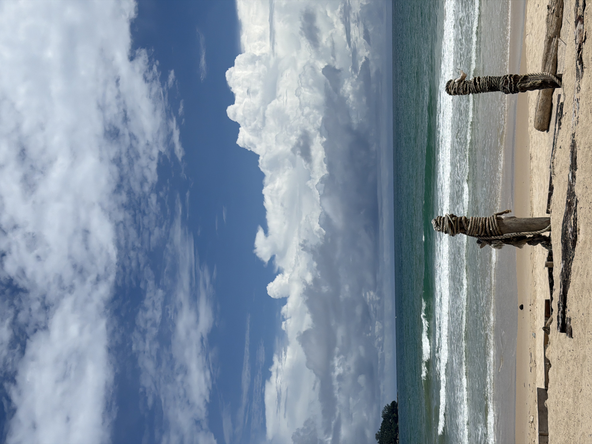 The image captures a picturesque beach scene on a sunny day, likely in Choeng Thale, Thailand. The composition is dominated by the vast expanse of the sky, filled with a dramatic array of clouds, ranging from wispy white streaks to large, fluffy cumulus formations. Below the sky, the turquoise ocean stretches out, with gentle waves breaking on the shore, creating white foamy lines that lead to the sandy beach.
In the foreground, the sandy beach is partially occupied by weathered wooden posts, wrapped with thick ropes. These appear to be remnants of some structure or possibly anchors for boats. A single bird is perched on the upper rope-wrapped post, adding a touch of life to the scene. The overall atmosphere is serene and tranquil, evoking a sense of peace and natural beauty. The time of day is likely daytime, with the sun illuminating the clouds and the water. The weather appears to be clear and pleasant, with the presence of both blue sky and significant cloud cover indicating a dynamic but fair weather condition. No text is visible in the image.