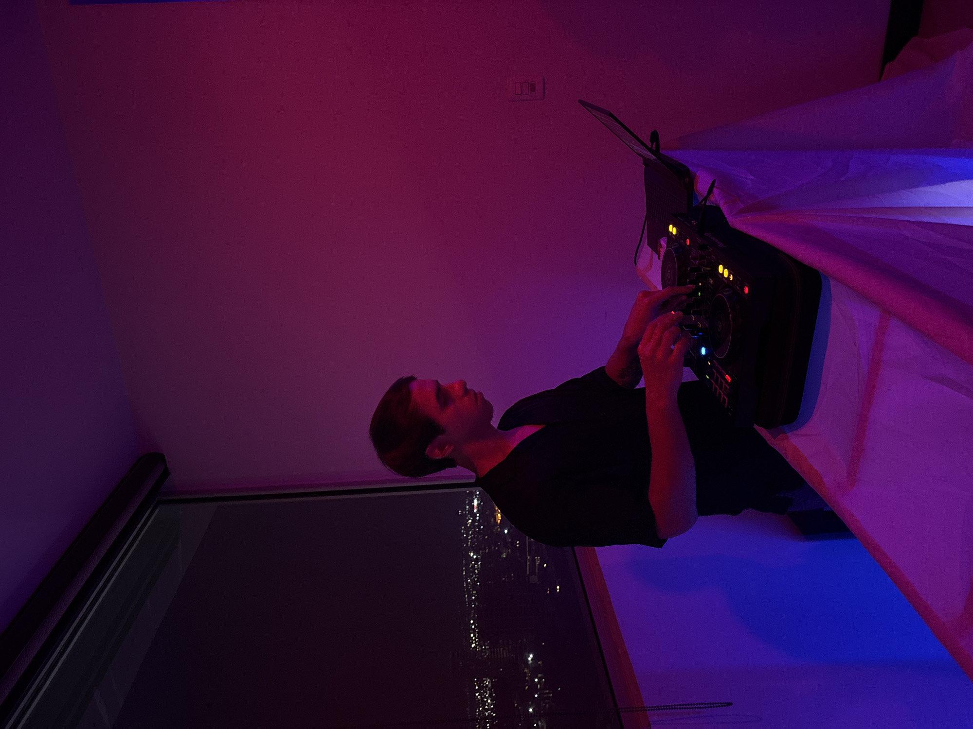 A man is standing in front of a DJ mixer in a room with pink and blue lighting. The background includes a large window showing a cityscape with numerous lights, presumably at night. A white cloth is draped over something in the upper right corner of the image. The man is wearing a black shirt. The location is San Bernardino Tlaxcalancingo, Mexico.