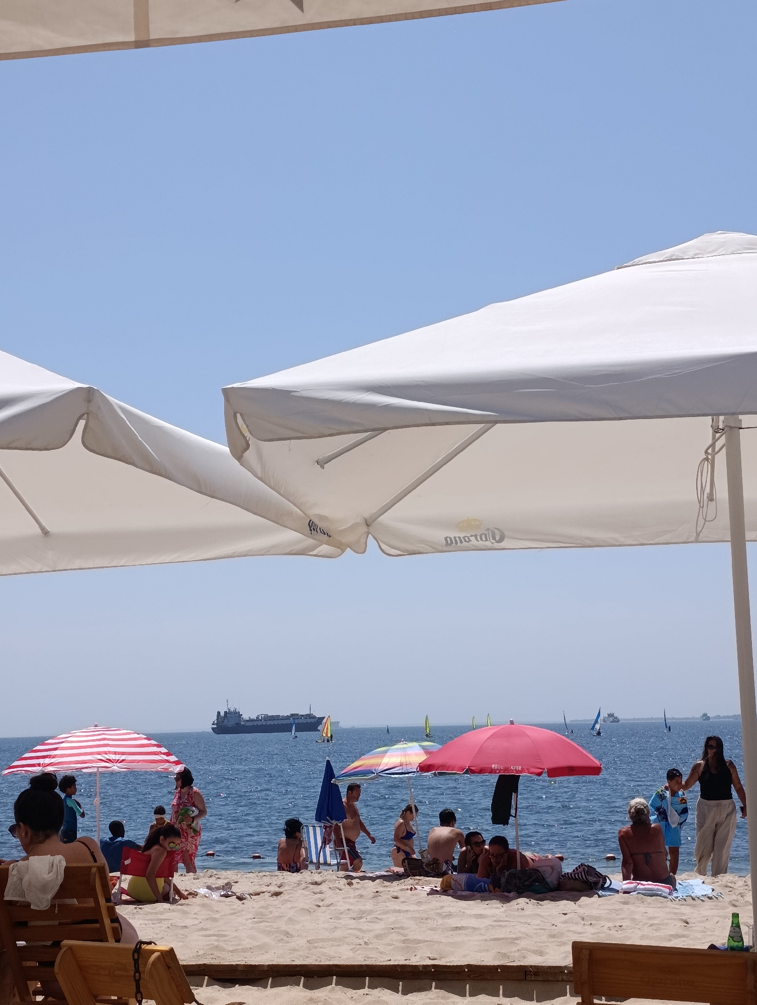 This is a photograph taken on a sunny day at a beach in Setúbal, Portugal. The foreground is dominated by a wooden beach chair and part of a wooden structure, suggesting a relaxed, recreational setting. In the midground, people are scattered across the sandy beach, some relaxing under umbrellas, others near the water's edge. A red and white striped umbrella shades a group of people, including children. A bright red umbrella provides shade for another group, with a person standing nearby in light-colored pants. A few other umbrellas, including a colorful one, are also visible. The sea stretches out in the background, dotted with several sailboats. A large cargo ship is visible on the horizon, indicating shipping activity in the area. The sky is clear and blue. The overall scene depicts a typical summer day at the beach, with a mix of relaxation and light activity.