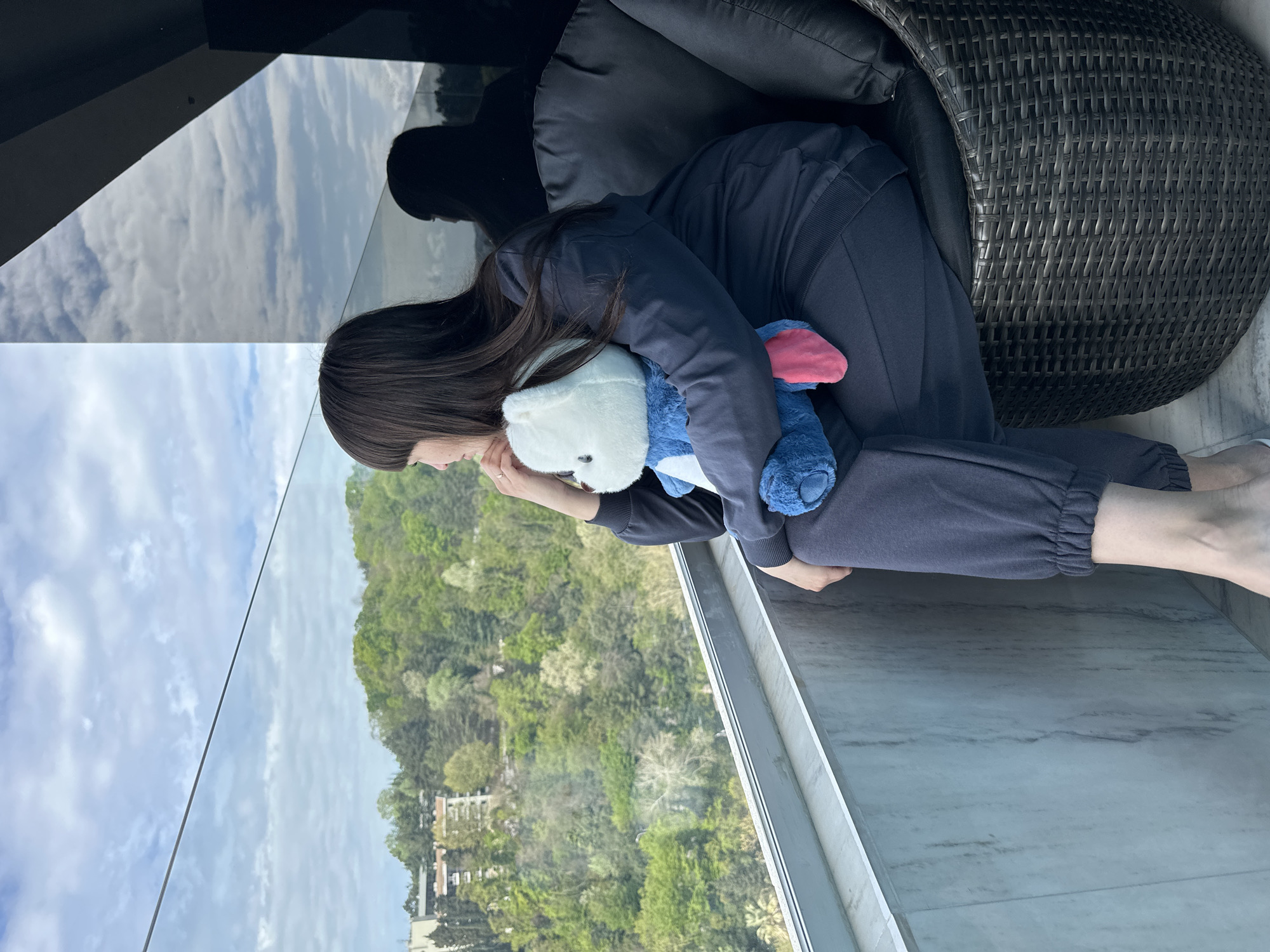 This vertically oriented image features a woman sitting on a modern balcony, gazing out at a lush, green landscape under a partly cloudy sky. The setting, identified as Sochi, Russia, showcases rolling hills and mountains densely covered with trees, with a few buildings scattered in the distance.
The woman has long, dark hair and is wearing a casual dark blue matching tracksuit. She is seated in a dark brown woven outdoor chair with dark cushions, positioned near a clear glass balcony railing. She holds two stuffed animals: a white, polar bear-like plush and a blue one with pink ears, resembling Stitch. Her posture suggests a moment of contemplation or relaxation as she takes in the panoramic view.
The sky above is bright with white clouds, which are visibly reflected in the dark, reflective overhang of the balcony ceiling and subtly in the glass railing. The balcony floor appears to be light-colored marble or stone. The scene is well-lit, indicating daytime. No text is visible in the image.