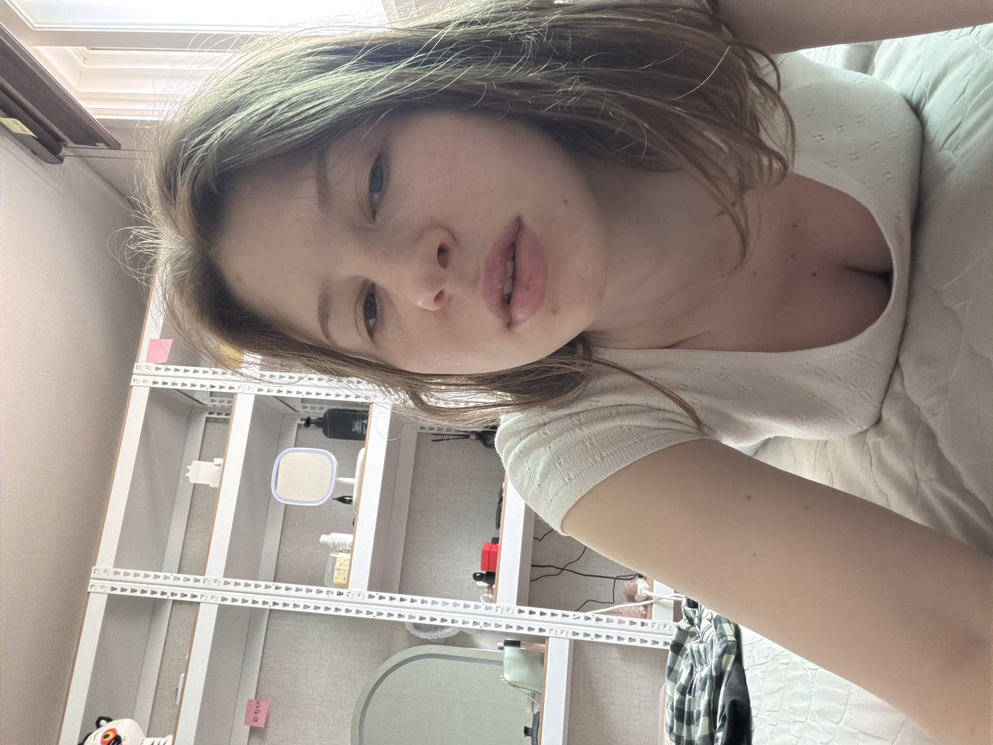 A young woman with shoulder-length brown hair is lying on a white, textured bed, with her body angled towards the camera. She is wearing a short-sleeved, white, textured top with a low neckline. Her head is tilted slightly, and she is looking directly at the camera with a neutral expression. Her face is the primary focus, with her eyes, nose, and lips clearly visible. In the background, a white metal shelf unit is visible, filled with various items including a stuffed animal, a pink sticky note, containers, bottles, a small mirror, and a black dispenser. Further to the left, part of a light green oval mirror and a small mint green purse are on a shelf. The environment appears to be an indoor bedroom. The image captures the woman from the chest up, with her arm extending towards the viewer. The lighting suggests it is daytime, with light coming from a window to the right of the frame.