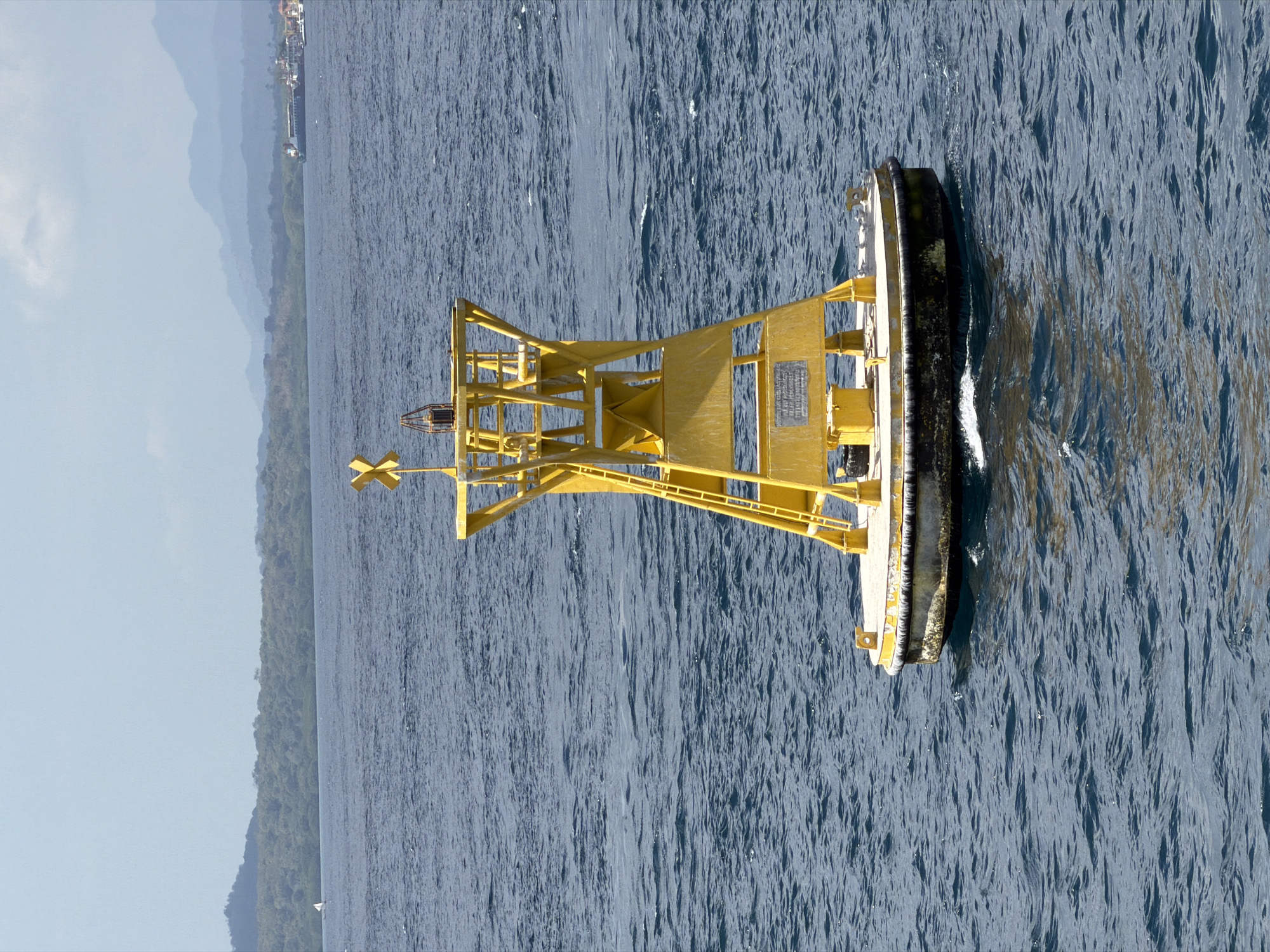 A bright yellow navigational buoy with a black fender is floating in choppy, dark blue water. The buoy has a tall, open-frame structure with a ladder leading up to a platform at the top, where a light fixture is visible. A yellow cross-shaped marker is attached to the top. The buoy appears to be in a harbor or channel, with a lush green, tree-covered coastline in the background under a hazy blue sky. In the distance, mountains are faintly visible, and a ship is present on the horizon. The scene is set in the city of Ketapang, Indonesia.