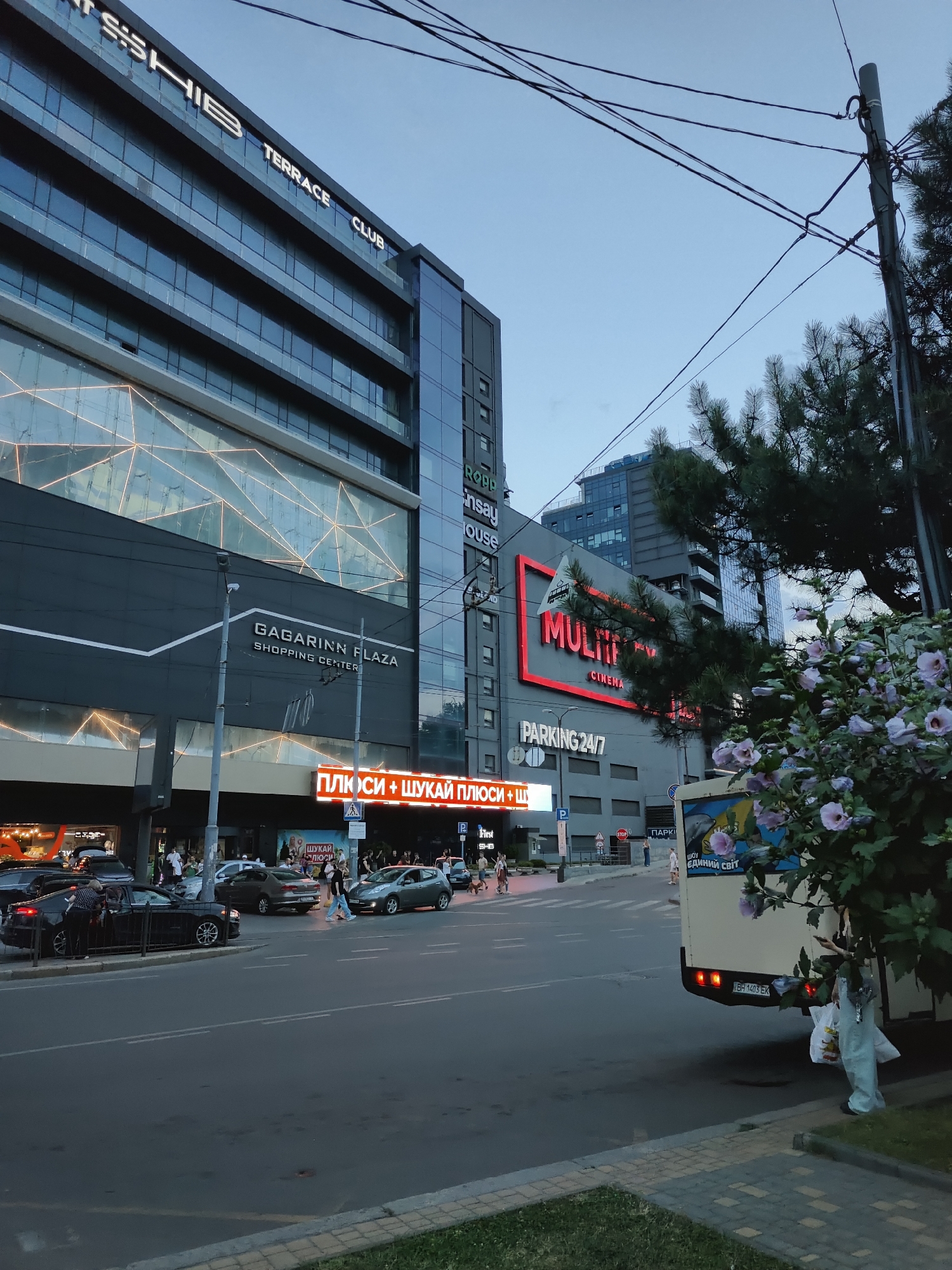 The image captures a street scene in Odesa, Ukraine, featuring the Gagarinn Plaza shopping center. In the foreground, a street with several cars is visible, including a black car being attended to by a person. Pedestrians are crossing the street in the middle ground, with the illuminated sign for "MULTIMEDIA CINEMA" and "PARKING 24/7" prominent on the building's facade. A light-colored bus is parked on the right side of the image. Lush green trees and flowering bushes add to the urban landscape, with utility poles and overhead wires indicating an active city environment. The sky is a muted blue, suggesting late afternoon or early evening. The architecture of the Gagarinn Plaza is modern, characterized by glass and dark-colored panels.