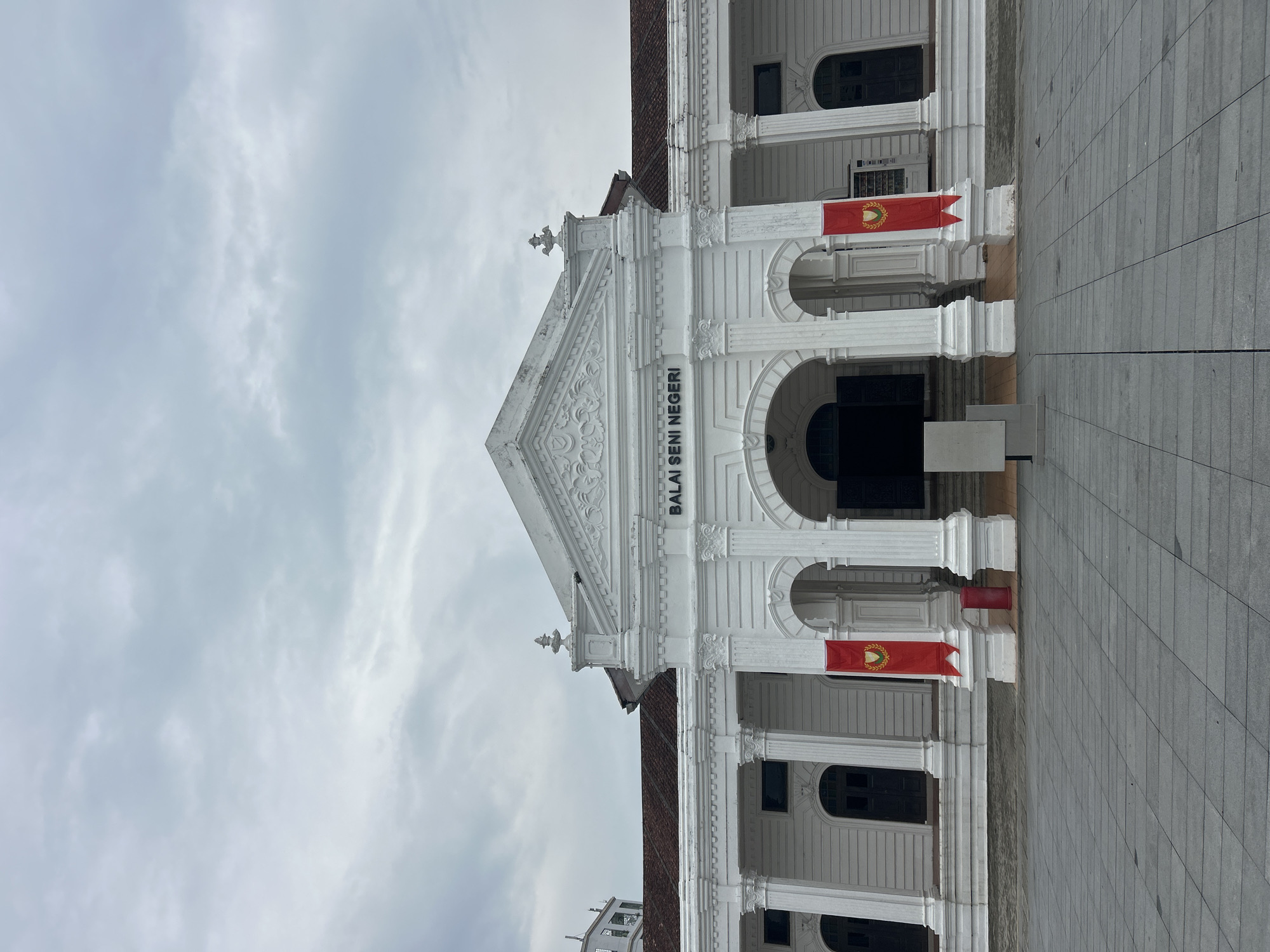 The image shows the facade of a white neoclassical building with a grand entrance. The building is identified by the text "BALAI SENI NEGERI" above the entrance, which translates to "State Art Gallery." Two red banners with a gold crest hang from columns flanking the entrance. The building has a symmetrical design with white columns and dark arched doorways. The sky is overcast with grey clouds. The foreground features a paved plaza with large square tiles. The building is located in Alor Setar, Malaysia, and is the State Art Gallery of Kedah.
