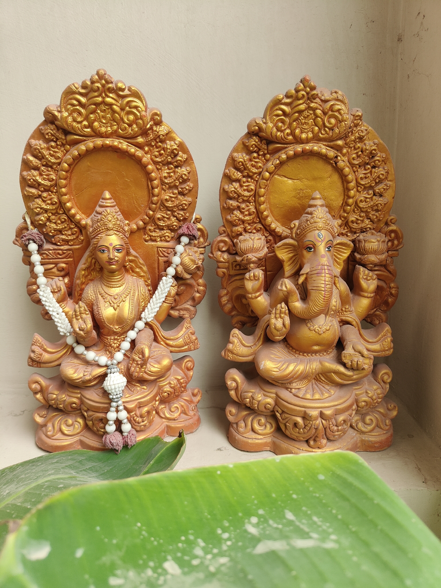 The image shows two golden statues, likely deities, placed side-by-side against a neutral, possibly beige or light brown, textured wall. The statue on the left depicts a female figure with multiple arms, adorned with a white beaded necklace. The statue on the right is of a Ganesha, identifiable by its elephantine head and multiple arms. Both statues are seated on ornate bases and have elaborate decorative backdrops. In the foreground, partially obscuring the lower part of the statues, are large, green leaves, suggesting an outdoor or semi-outdoor setting. The lighting appears to be natural, casting soft shadows. The environment appears to be a domestic or religious setting. The presence of these deities suggests the location is likely in India, possibly in Gurgaon, given the request, which is a major city in Haryana, India, known for its Hindu population and numerous temples.