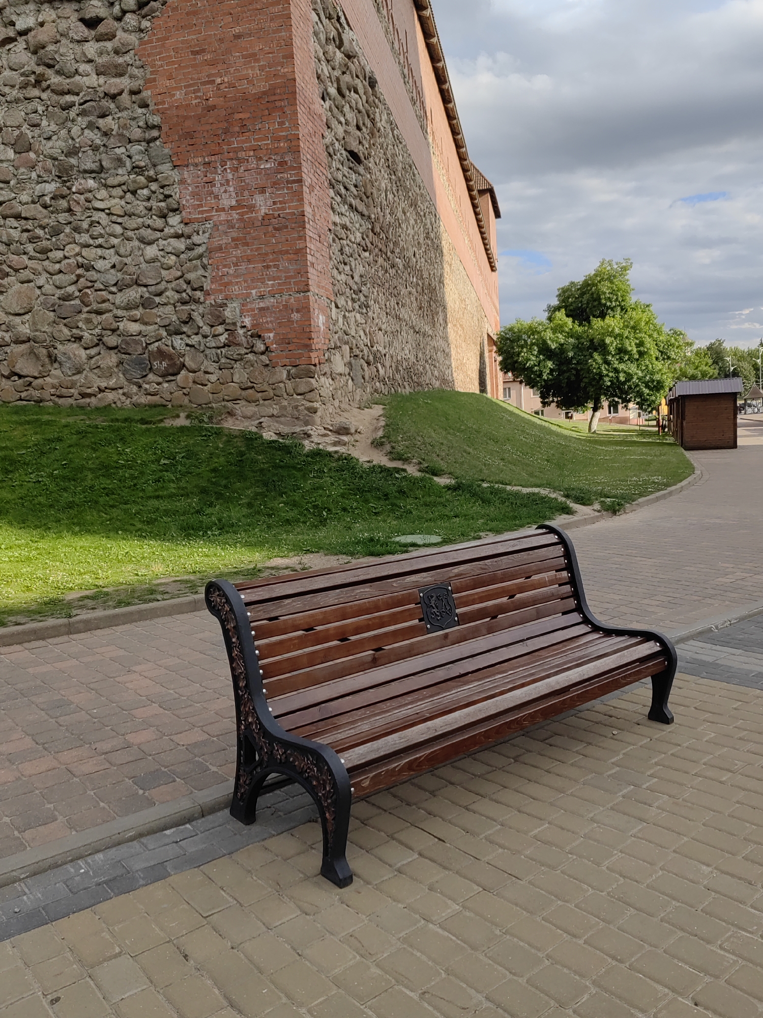 An outdoor scene features a wooden park bench in the foreground, positioned on a paved walkway. The bench is made of dark wood slats and has ornate black metal legs. Behind the bench, a grassy slope leads up to a large, weathered brick and stone wall, which appears to be part of a historical structure. Further back, a green tree stands to the right of the wall, and beyond that, a small wooden shed and a paved area are visible. The sky above is partly cloudy with patches of blue. The overall setting suggests a public space with a historical element.