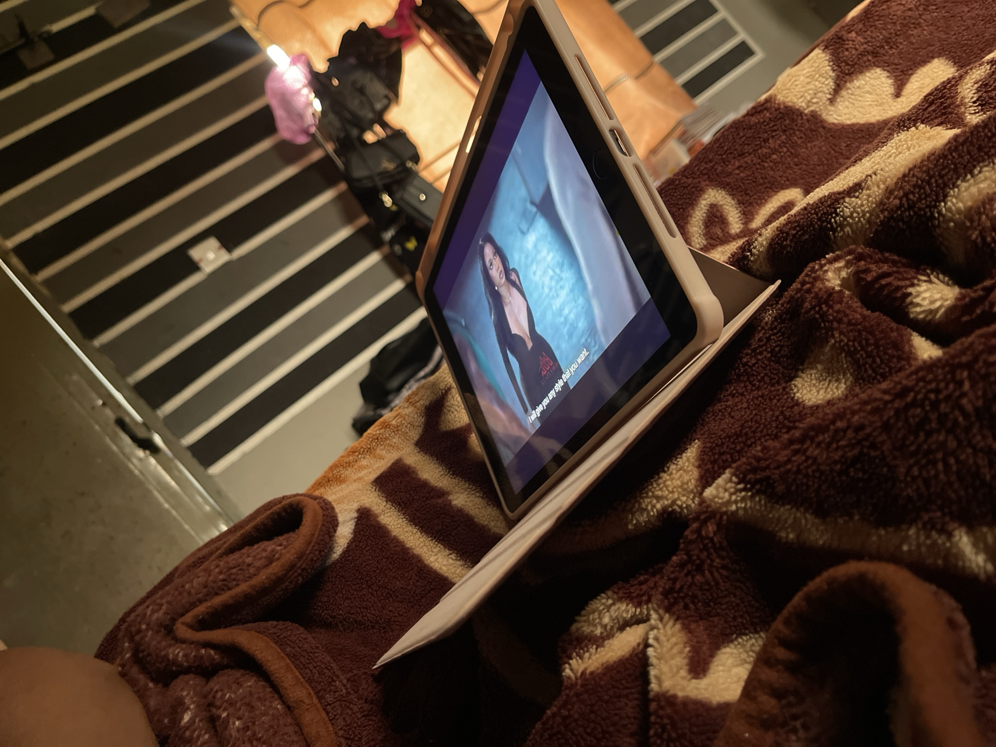 An indoor scene at night shows a tablet computer propped up on a patterned blanket, displaying a woman in a black dress. The tablet is held at an angle, and its screen is partially visible. The background is out of focus, but appears to be a room with striped walls and a light source. The overall impression is that of a casual, private setting, likely a bedroom or living area. The specific location in Rafin Par, Nigeria, cannot be determined from the visual information provided in the image.
