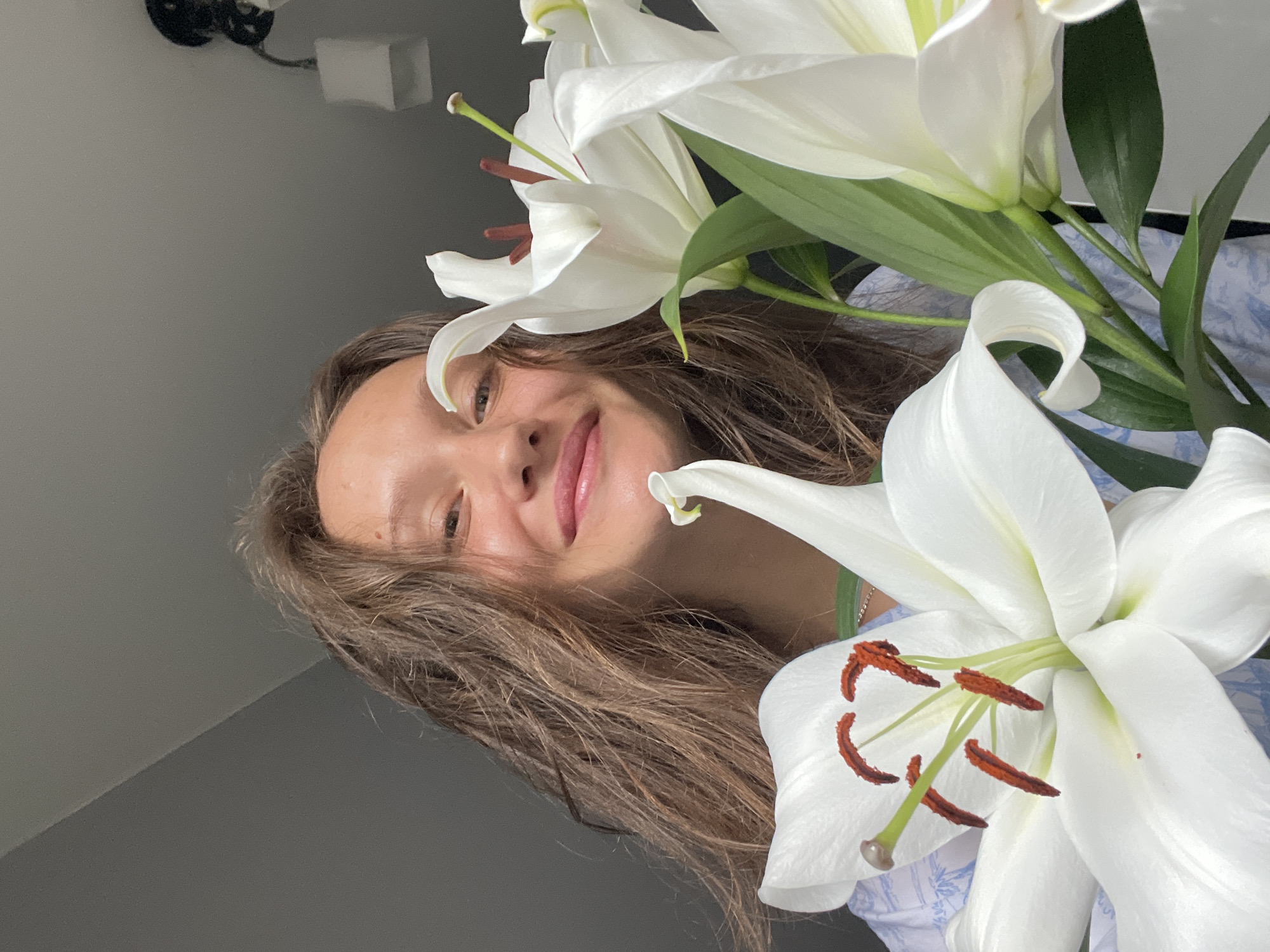 An indoor, eye-level to slightly low-angle close-up depicts a smiling young adult female positioned behind a bouquet of white lilies. The female, centered in the upper portion of the frame, possesses medium-length brown hair, a fair complexion, and a visible mole on her upper forehead. She wears a light-colored garment with a subtle patterned design, possibly light blue, and a thin silver-colored chain around her neck. Her expression is a closed-mouth smile, and she is looking directly into the camera.
Prominently displayed in the foreground and extending through the lower and right portions of the image are multiple large white lily blooms. These flowers exhibit white petals, visible green stems, dark reddish-brown anthers, and green pistils. Green foliage is also present among the flower stems.
The background consists of an interior space. The upper left area shows a plain grey wall transitioning into a lighter grey ceiling. The upper right segment reveals a ceiling, possibly white or very light grey, with a dark-colored light fixture featuring a white shade. The specific location within the city of Sochi, Russia, or any other geographic location, cannot be determined from the provided visual information. The overall lighting is even, suggesting an adequately lit indoor environment.