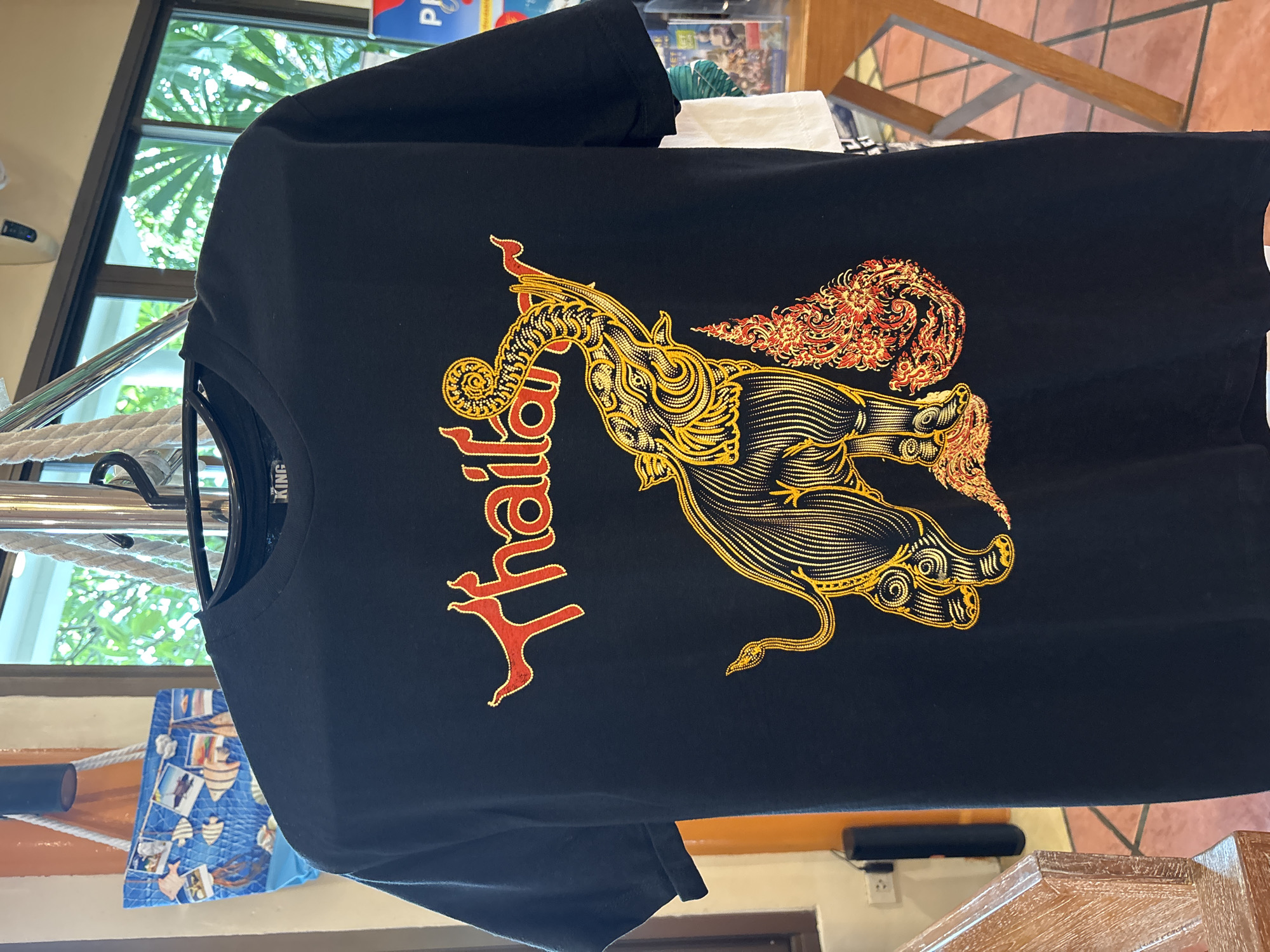 A black t-shirt with a graphic design is hanging on a hanger in what appears to be a shop in Chalong, Thailand. The graphic features the word "Thailand" in red and yellow lettering above an ornate depiction of an elephant. The elephant is outlined in yellow with intricate patterns, and it is standing next to a decorative element that resembles flames or elaborate foliage, also in red and yellow. The t-shirt has short sleeves and a crew neck. In the background, a window reveals lush green foliage, suggesting an outdoor or tropical setting. A blue tote bag with a nautical-themed print, featuring fish and boats, is visible to the left of the t-shirt. Various shelves and wooden furniture are also present in the shop.