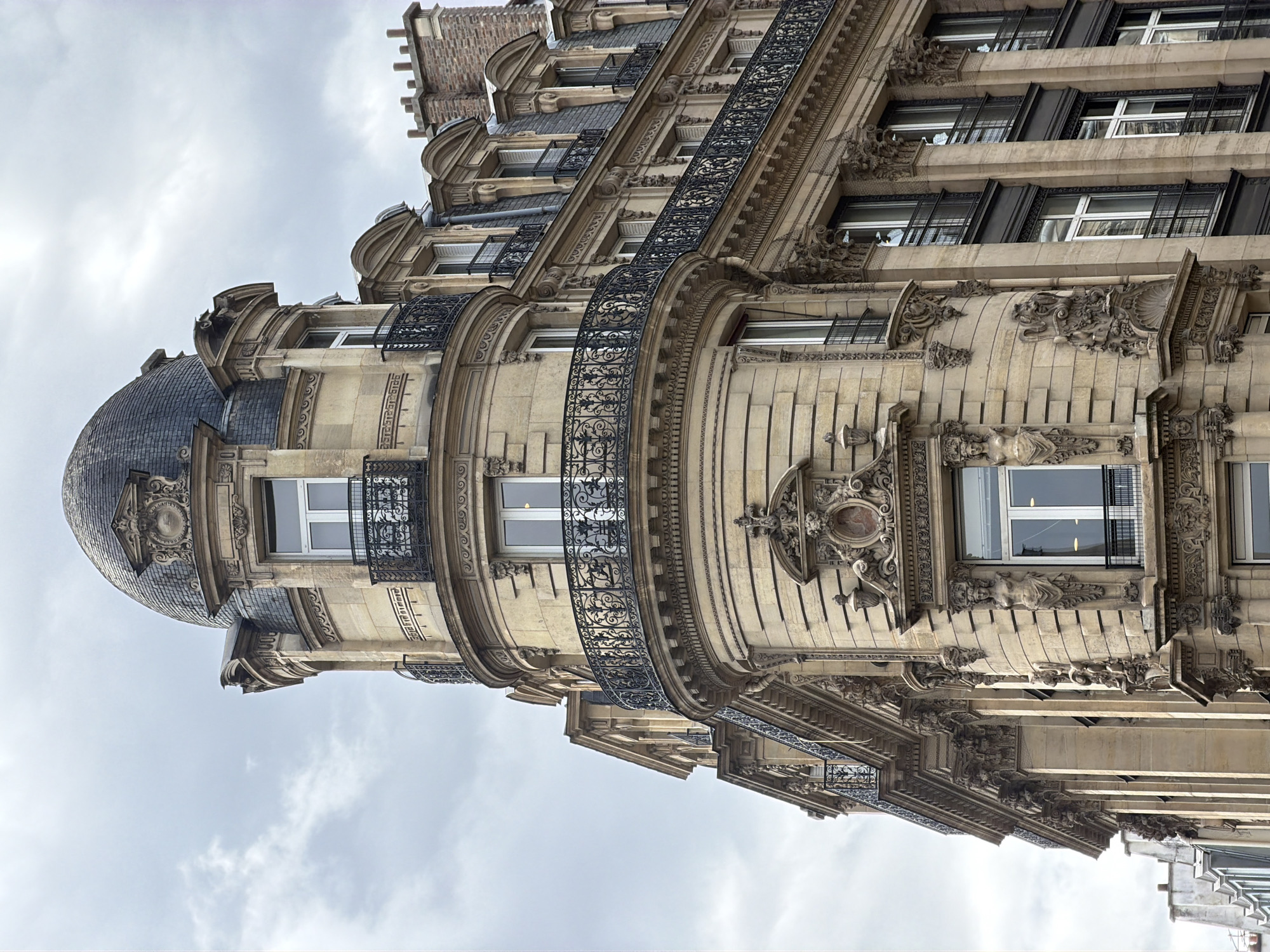 The image depicts a corner of a building in Paris, France. The architectural style is ornate, with elaborate moldings, carvings, and wrought-iron balconies. The building is constructed of light-colored stone. The corner of the building features a rounded turret-like structure topped with a dome covered in dark tiles. Several windows are visible, some with dark shutters. The sky is overcast with white clouds. No people or animals are visible in the frame.