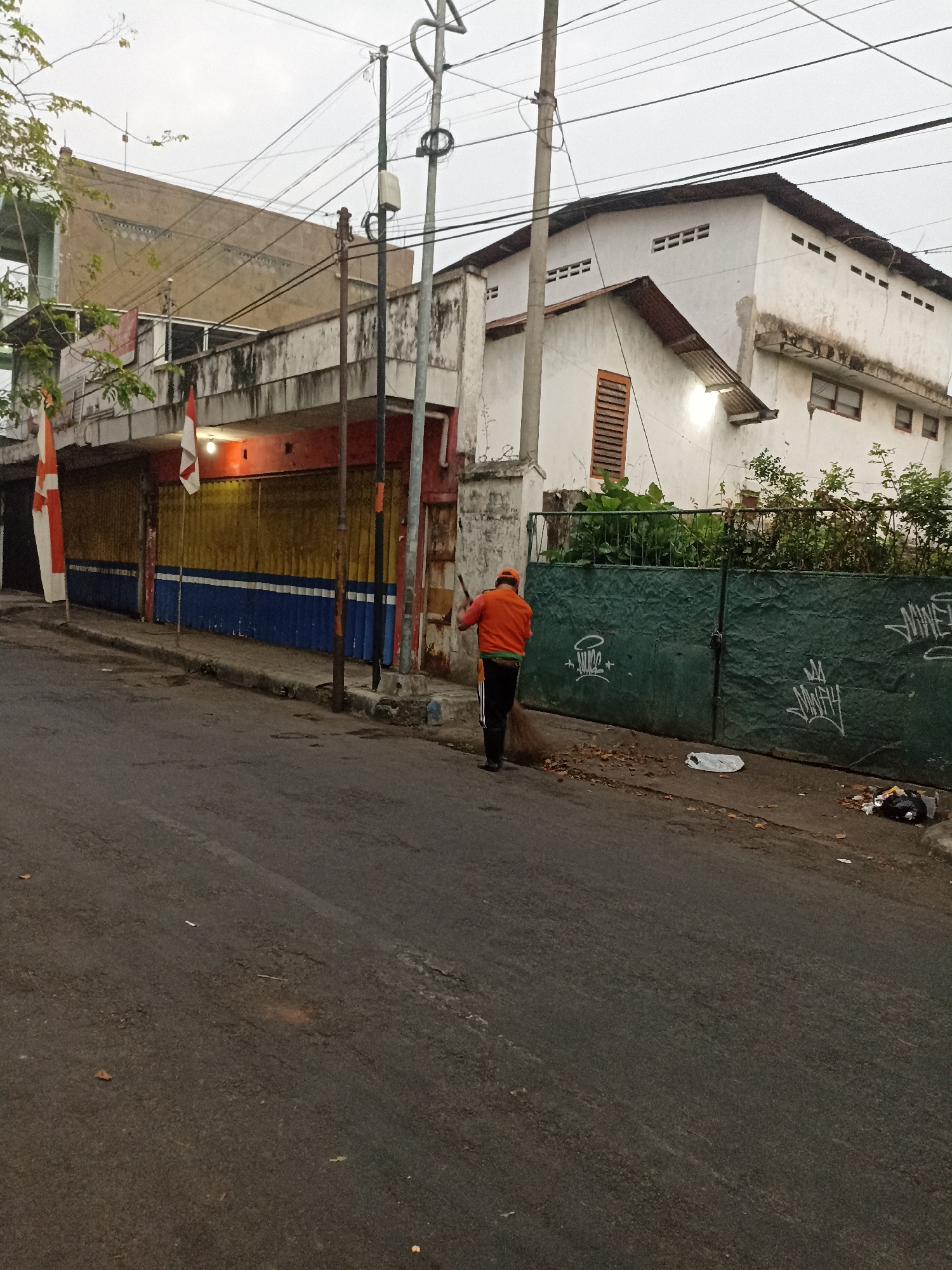 A person wearing an orange jacket and a baseball cap is sweeping a paved road with a broom in Lumajang, Indonesia. The individual is facing away from the camera, moving from right to left, and appears to be gathering fallen leaves and debris. To the left of the person, there is a building with a facade painted in horizontal bands of blue, yellow, and red, with two Indonesian flags displayed. Further to the left, a multi-story building with a cream-colored exterior and a balcony is visible. To the right of the sweeper, a tall green fence with graffiti adorns its surface, with overgrown vegetation behind it. A white plastic bag and trash are scattered on the ground near the fence. The sky is overcast, and the street appears to be a commercial or residential area.