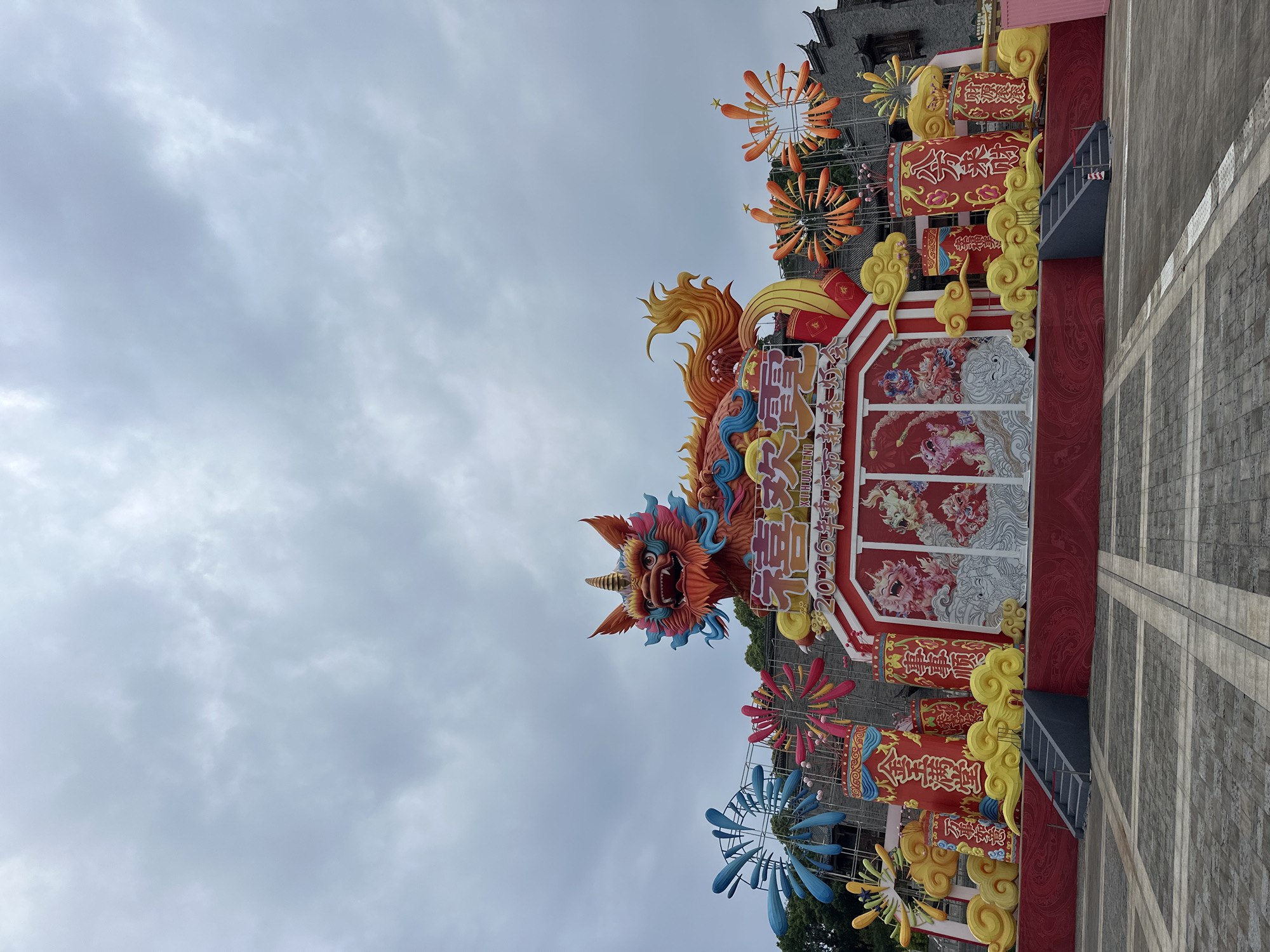 The image shows a large, elaborate outdoor display, possibly for a festival or celebration. Dominating the display is a large, colorful dragon-like figure with a fierce expression and ornate details. Below the dragon, there are decorative panels with images and Chinese characters. The characters and imagery suggest themes of prosperity, happiness, and good fortune. There are also structures decorated with vibrant colors, traditional Chinese patterns, and decorative cloud motifs. Above the dragon, there are orange floral pinwheels. The foreground features a paved area with concrete tiles.
The text on the panels includes Chinese characters translating to wishes such as "Prosperity", "All Wishes Come True", "Wealth Comes from All Directions," and "Happy Life". There is also a mention of the "Chongqing Liangjiang 2026 New Year Lantern Festival," indicating that the display is likely part of this event in Liangjiang, China.