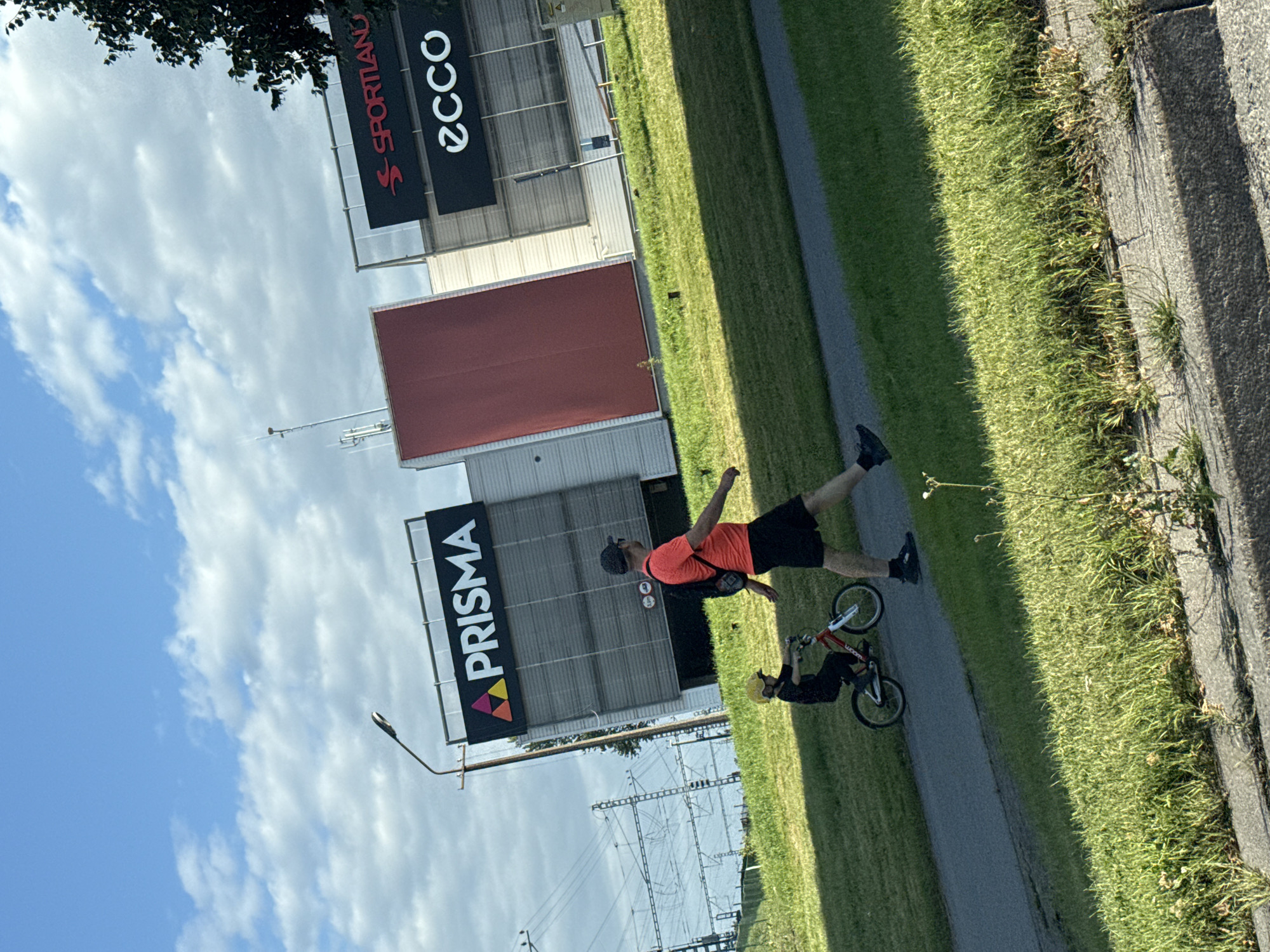 The image shows a man and a child outdoors on a sunny day, with a building in the background. The man, wearing an orange t-shirt and black shorts, is walking on a paved path, while the child, wearing a yellow helmet and dark clothing, is riding a red bicycle beside him. Behind them, a modern building with large signs is visible. One sign reads "PRISMA" in large white letters, and another displays the logo for "SPORTLAND" and "ecco". The surrounding environment includes a grassy slope, trees, a street light, and power lines, suggesting an urban or suburban setting. The sky is blue with scattered clouds.