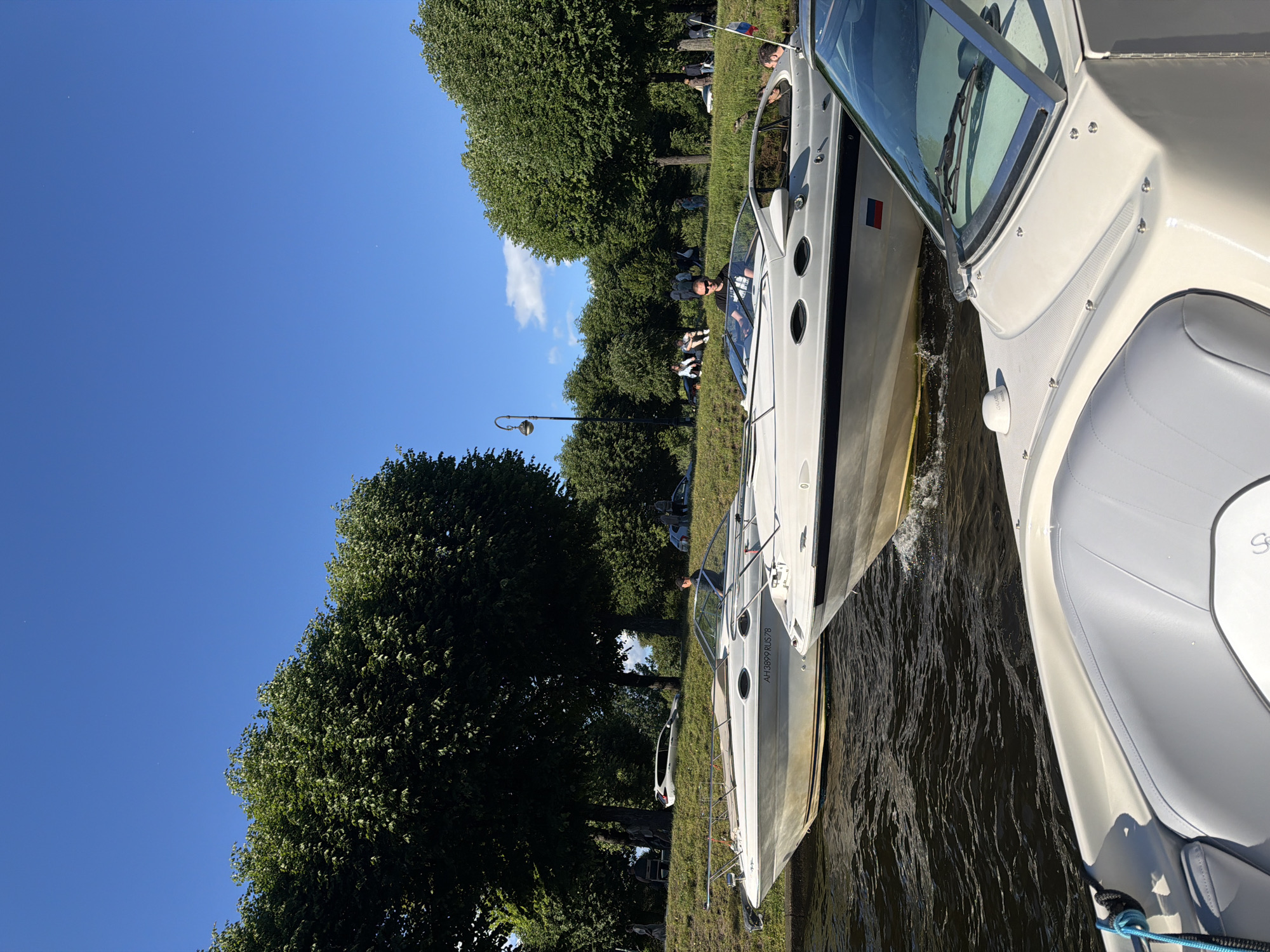This is a medium shot of two motorboats moored on a body of water, likely a canal or river, in Saint Petersburg, Russia. The boats are white and appear to be leisure craft. The scene is set on a sunny day with a clear blue sky and some fluffy white clouds. Lush green trees line the grassy bank behind the boats, suggesting a park or waterfront area.
On the bank, several people are visible, some sitting and others standing, enjoying the day. One person is seated in the cockpit of the boat in the foreground, and another is standing at the helm of the boat in the background, appearing to be operating it. A blue rope is tied to the bow of the foreground boat.
Notable details include the reflection of the boats on the water, the dappled sunlight filtering through the trees, and a lamppost on the bank. The overall mood is relaxed and leisurely, indicative of a pleasant summer day. The visible text on the stern of the boat in the background reads "AH 3859 RUS 78", which is likely its registration number, and implies the location is Russia. The time of day appears to be late morning or early afternoon due to the bright sunlight and the angle of the shadows.