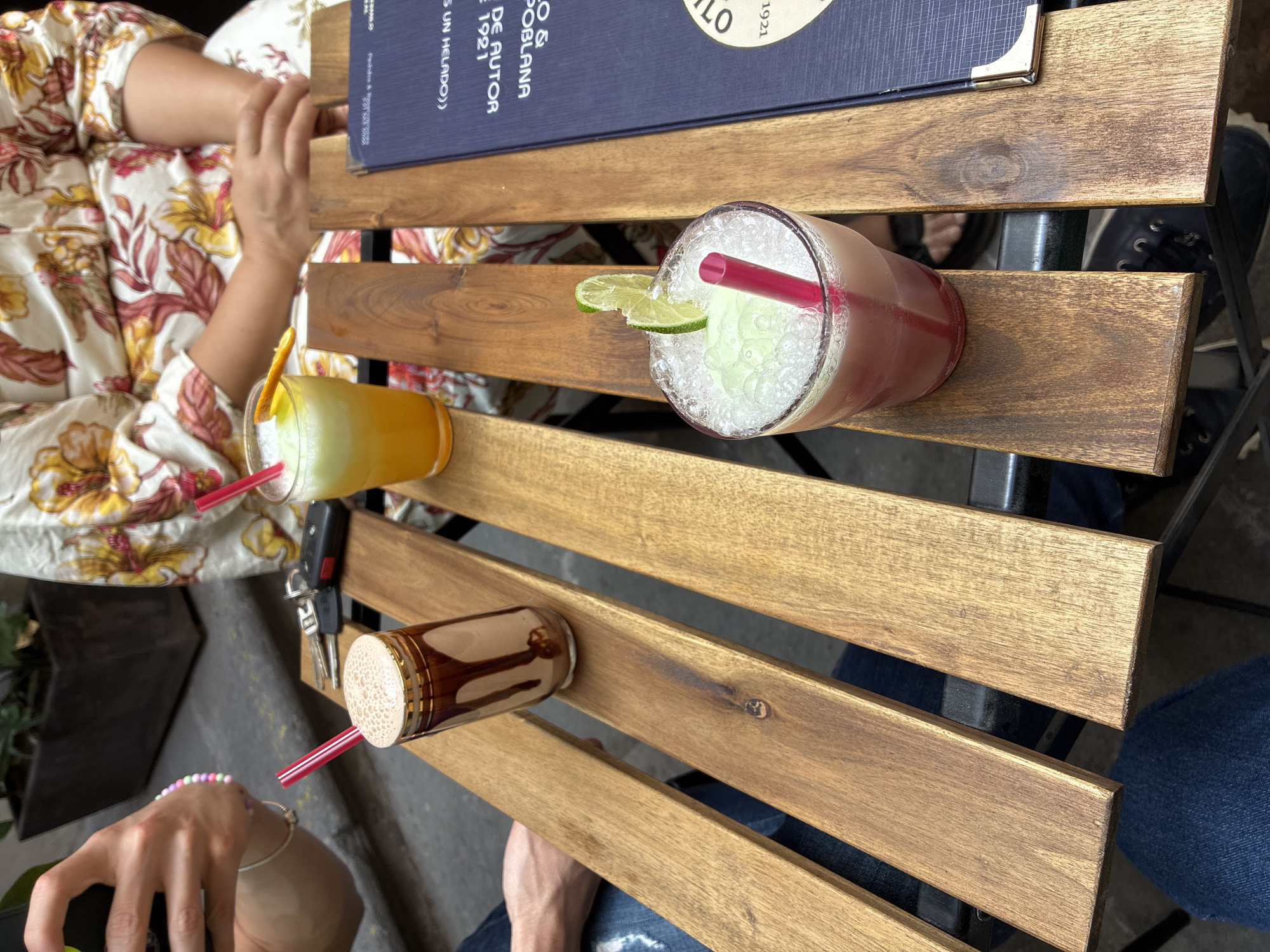 The image shows a wooden slat table with three beverages. From left to right: an orange-colored drink with a red straw, a pink drink with ice and a lime garnish and a red straw, and a brown and white beverage with a red straw. A blue menu lies on the table next to the pink drink. Parts of two people are visible; a person in a floral print shirt is partially visible at the top left, and a person in blue jeans is partially visible on the right. Keys sit next to the orange drink. A person wearing a beaded bracelet is visible at the bottom left. The image suggests an outdoor cafe setting. The writing on the menu indicates the location is in Puebla, Mexico.