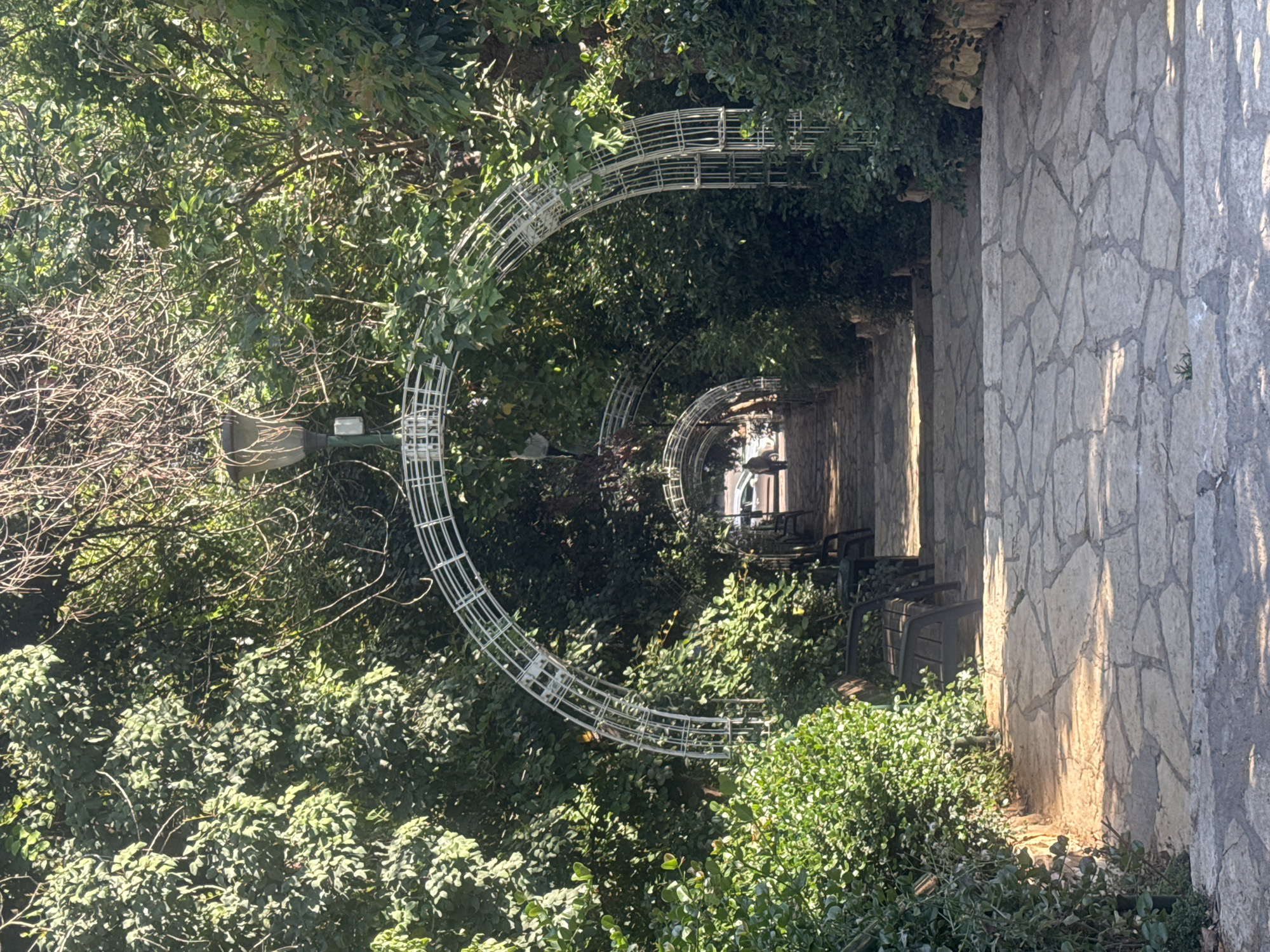 The image presents a pathway in Ramat Gan, Israel, defined by stone walls on one side and dense foliage on the other. Overarching the pathway are metal arches, creating a tunnel-like effect. The path itself appears to be paved. In the distance, chairs are visible along the wall. A lamppost stands amidst the greenery on the left. The overall impression is that of a secluded garden path.