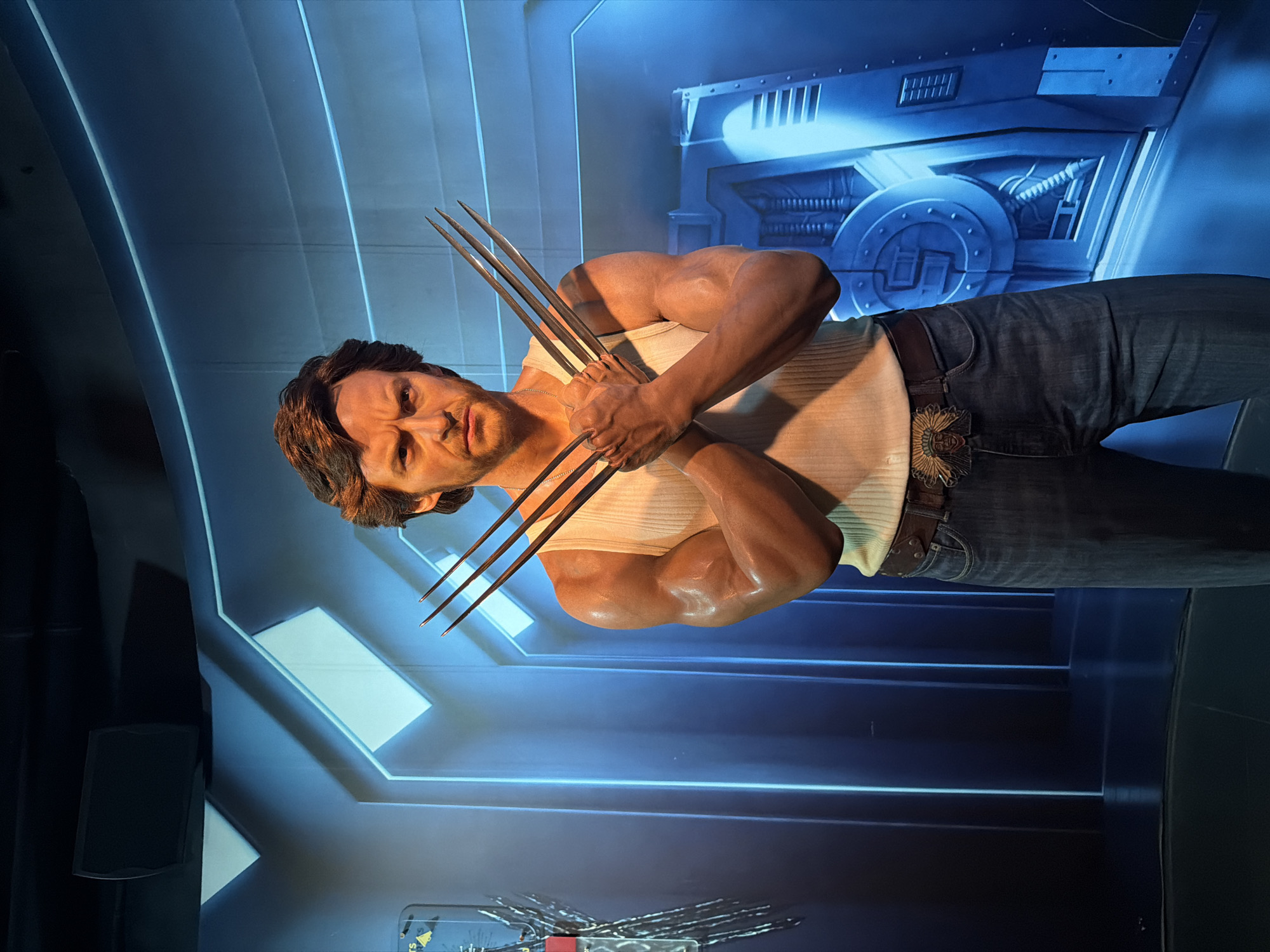 A wax figure of Wolverine, a fictional Marvel Comics character, stands with its arms crossed in front of its chest, displaying adamantium claws. The figure is wearing a white ribbed tank top, a brown belt with a large buckle featuring an Native American chief, and blue denim jeans. The backdrop is a futuristic, blue-lit environment with glowing lines and a large, circular mechanical structure on the right. The figure is positioned centrally in the frame, with its gaze directed forward. The setting appears to be an indoor exhibition or attraction, possibly a wax museum, and the overall ambiance is dark with dramatic lighting. The figure is highly realistic and detailed, with visible musculature and textured skin. The scene is static and posed. No specific location in Bangkok, Thailand, is discernible from the image itself.