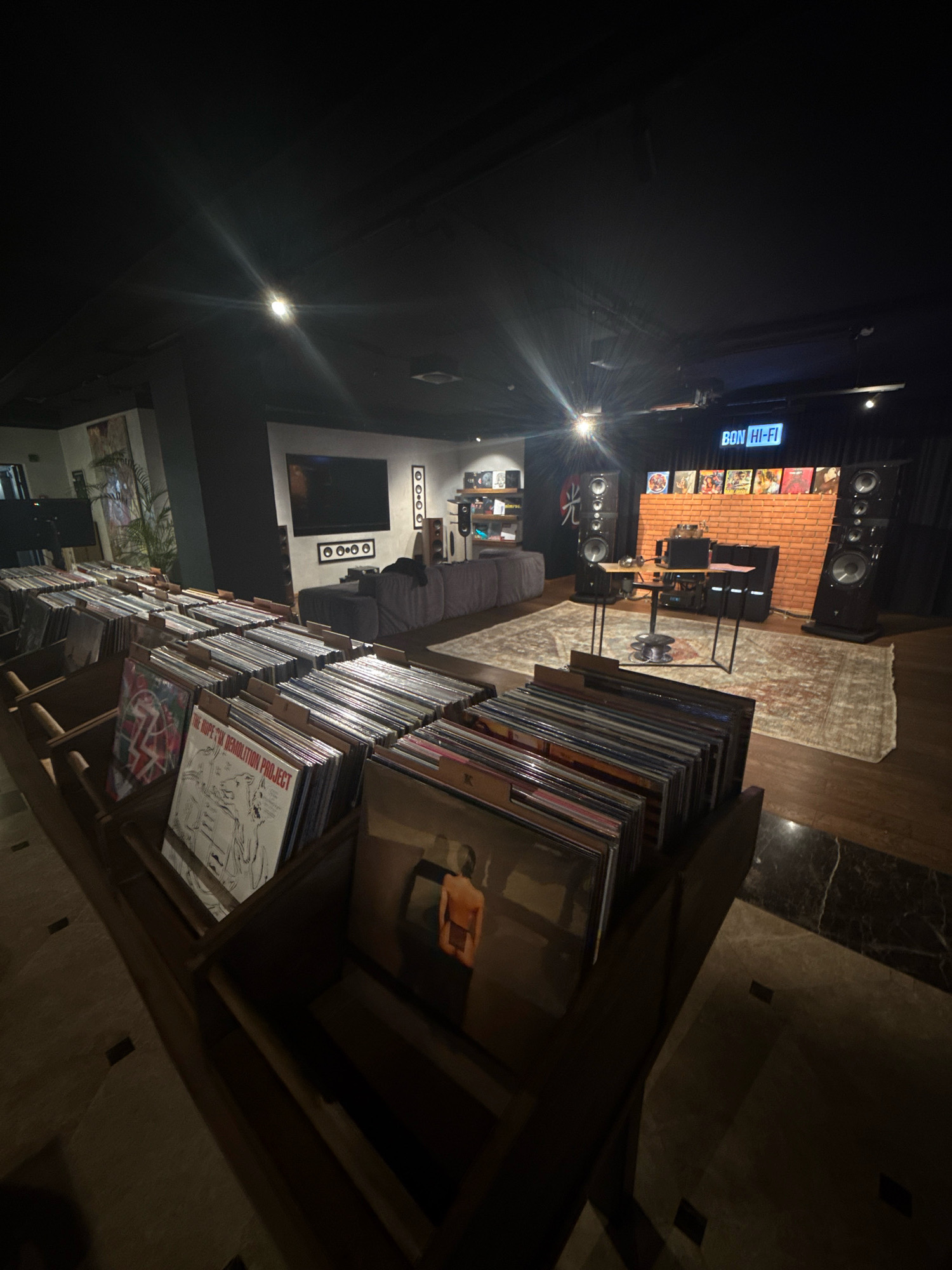 A dimly lit interior space, possibly a retail store or listening lounge for records, displays numerous rows of vinyl records organized in wooden crates. In the background, a brick wall is visible with a neon sign reading "BON HI-FI". Several large speakers are positioned around the room, and a rug with a decorative pattern covers a portion of the wooden floor. The overall impression is of a dedicated space for music enthusiasts. There are no people visible in the image. The location of Moscow, Russia is not ascertainable from the provided image.