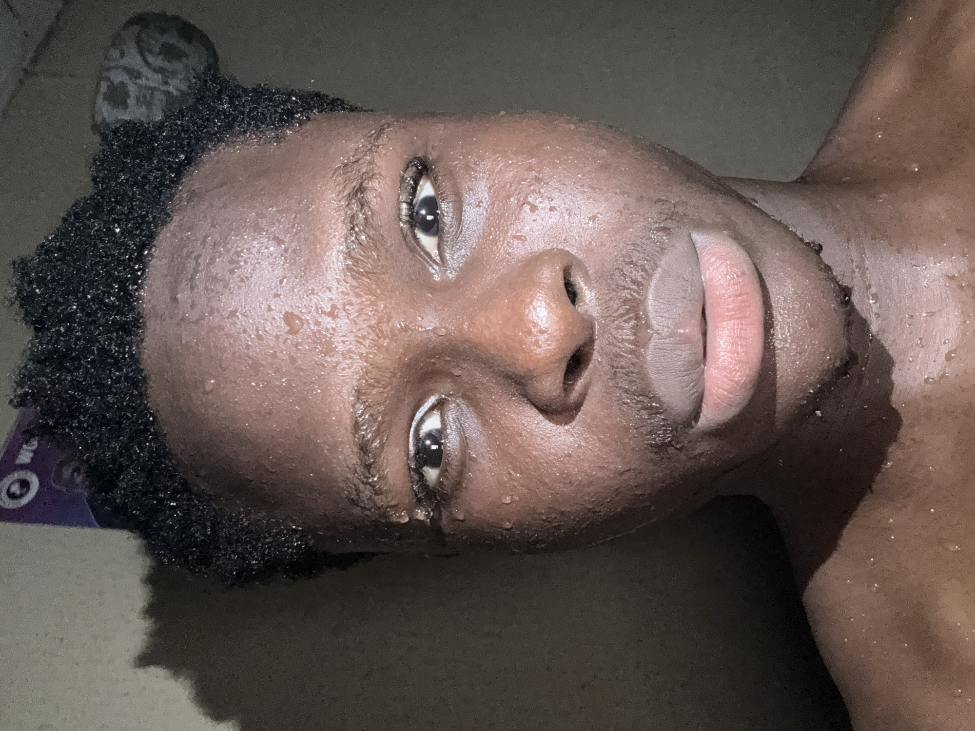 A close-up, dimly lit, slightly angled shot depicts the face and upper chest of a dark-skinned person with short, dark, curly hair. The skin appears damp with droplets of moisture. The subject gazes directly at the camera. The person's mouth is slightly open. A purple and white object is partially visible near the hair. The background is dark and indistinct. There is no indication of the location being Onitsha, Nigeria.