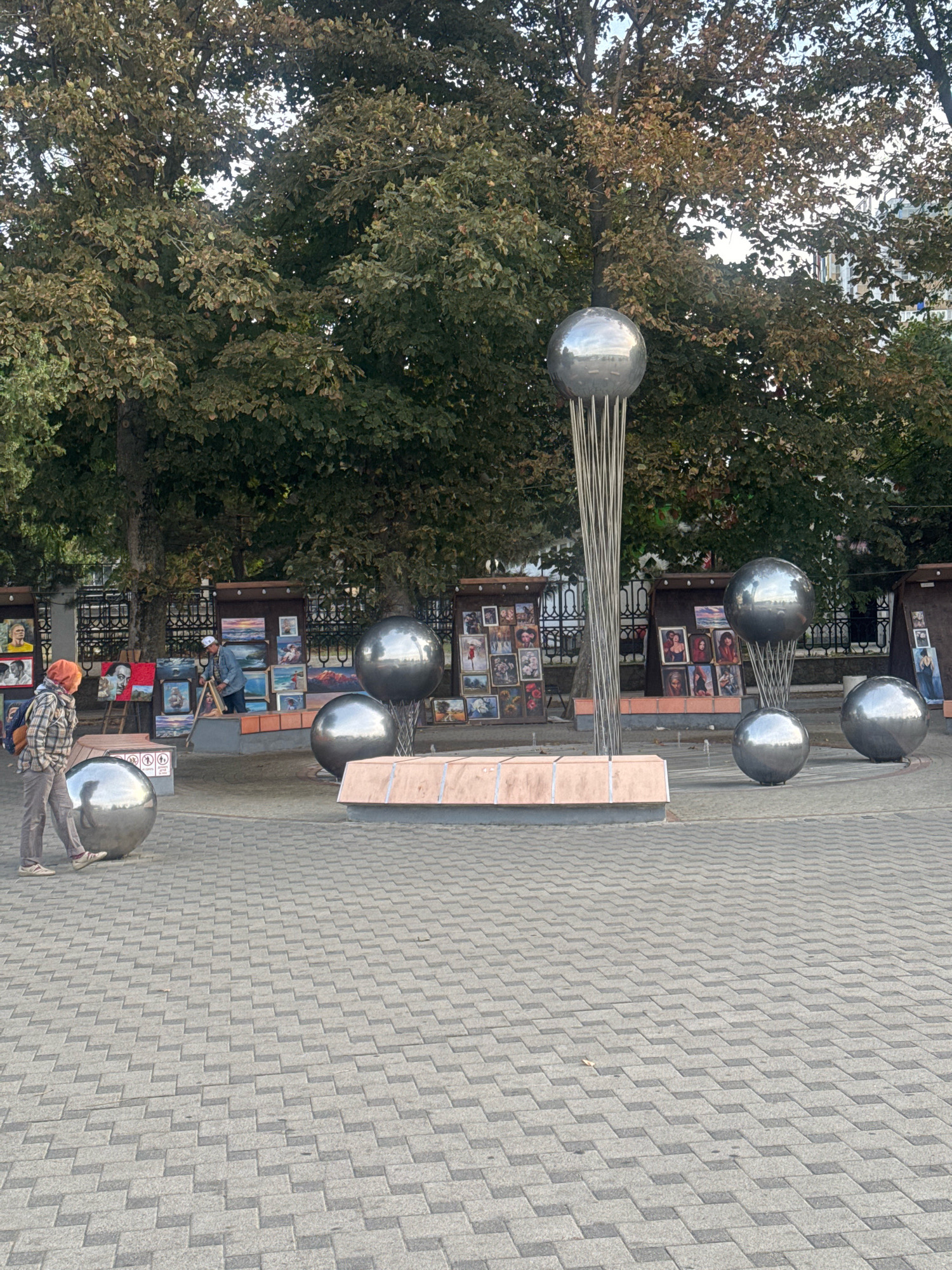 In Anapa, Russia, an outdoor art exhibition is displayed on a paved plaza. The scene is dominated by several large, spherical metal sculptures, with one in the center featuring a cluster of thin metal rods supporting a sphere. Behind the sculptures, art stands display numerous paintings, predominantly portraits and landscapes. Two individuals are present; one is a person wearing a plaid jacket and an orange headscarf, standing near a large metal sphere on the left. The second person, dressed in a blue shirt and a cap, appears to be an artist interacting with the artworks. The background is filled with mature trees, their leaves displaying autumnal colors, and a glimpse of buildings can be seen through the foliage. The ground is paved with interlocking grey bricks arranged in a geometric pattern.