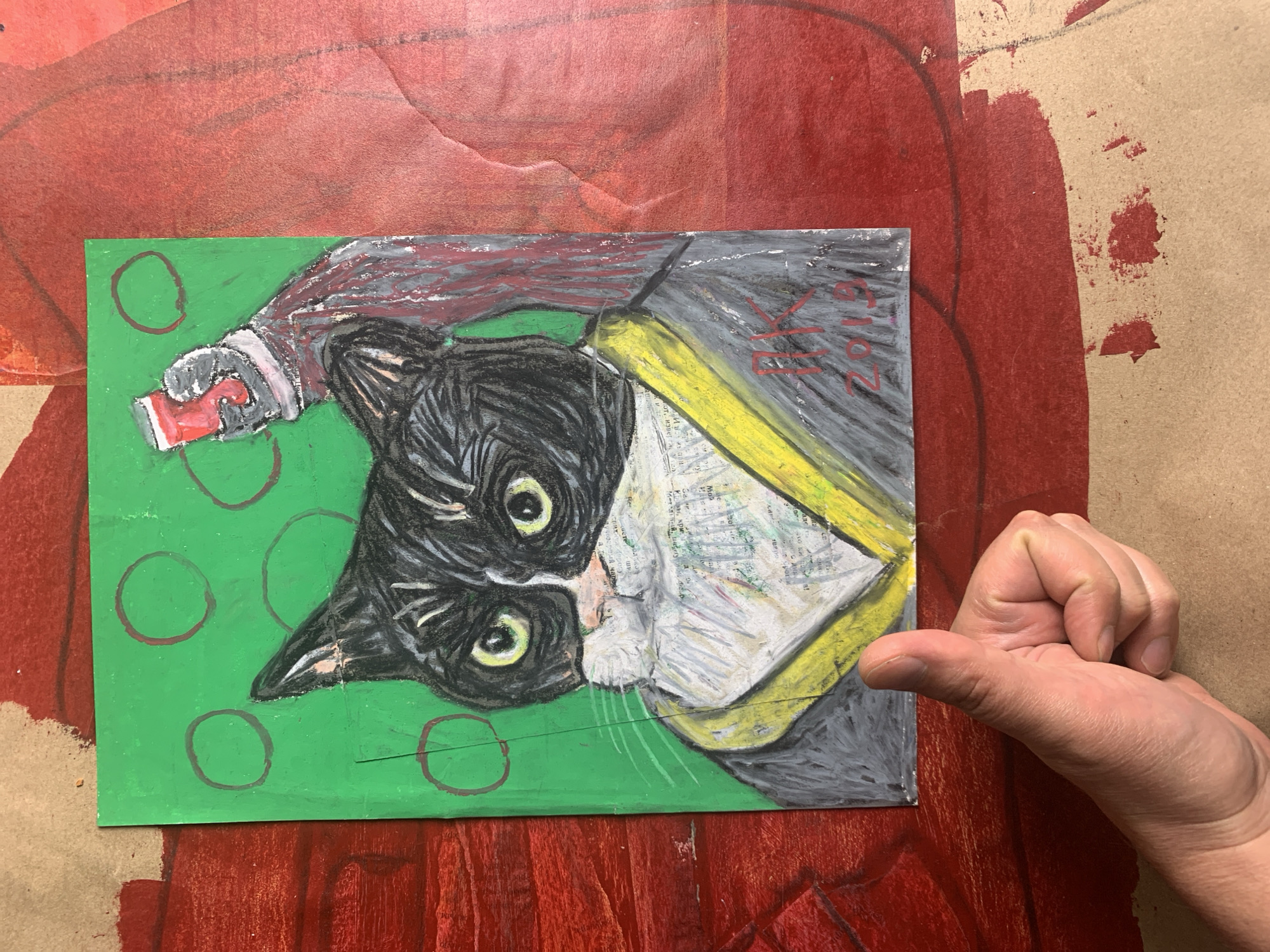 A close-up overhead shot shows a drawing of a black and white cat wearing a robe and holding a red cup in its paw. The drawing is on a green background with red circles. The drawing is signed "IK 2019". A hand gives a thumbs-up in the bottom left corner of the image. The background appears to be a red textured surface. There is no visible indication of the location in the city of Sergeevo, Russia.