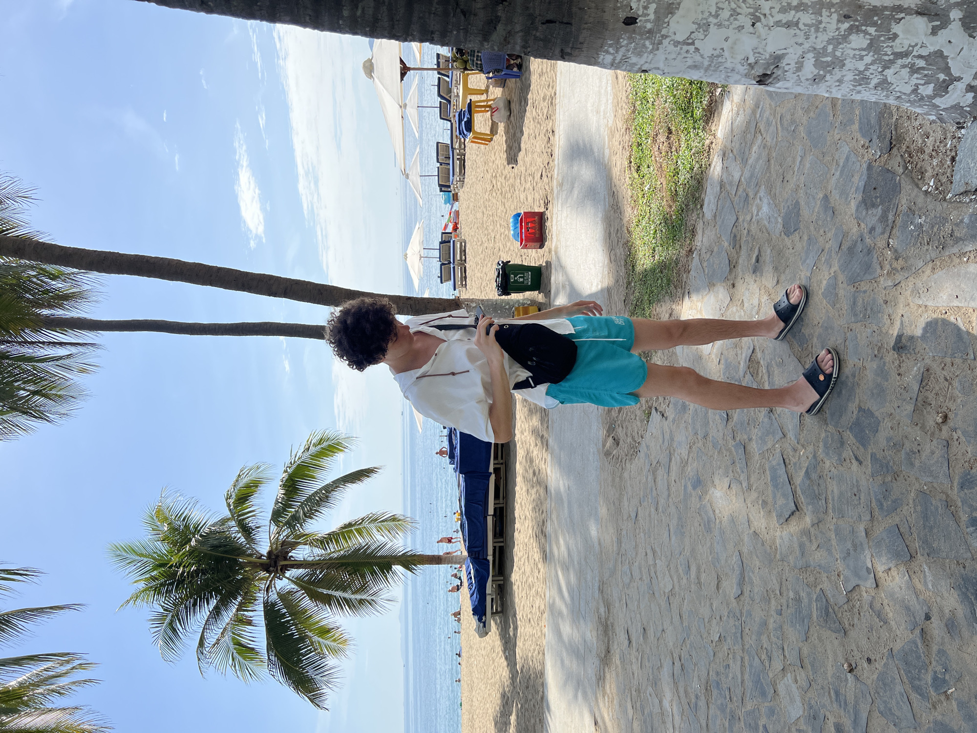 A young man with curly dark hair stands on a paved walkway on a sunny day at a beach in Nha Trang, Vietnam. He is wearing a loose-fitting white short-sleeved shirt, bright turquoise swim trunks, and black slide sandals. A black crossbody bag is slung over his left shoulder. He is looking down at something in his hands.
Behind him, a sandy beach is visible with rows of blue lounge chairs covered in dark blue fabric and white umbrellas. The ocean stretches out to the horizon under a clear blue sky with a few wispy clouds. Palm trees line the edge of the beach, their fronds casting shadows on the sand. Two trash cans, one green and one red, are situated on the sand near the walkway. In the distance, several people are visible on the beach and in the water. The scene conveys a relaxed tropical atmosphere.