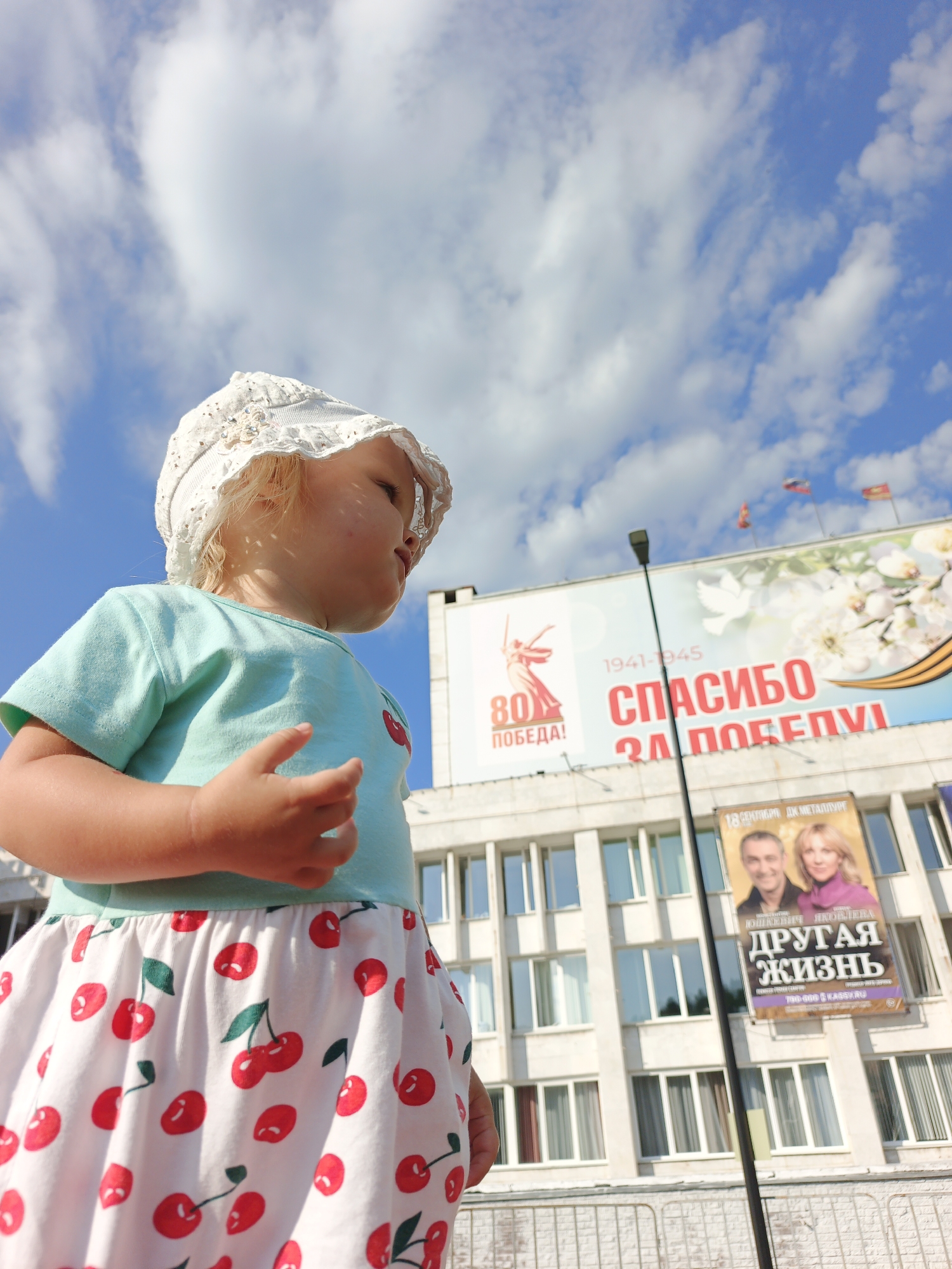 A low-angle outdoor shot features a young child wearing a white bonnet and a light blue t-shirt adorned with a cherry graphic. The child’s skirt is white with a pattern of red cherries and green leaves. The child is positioned in front of a light-colored building with multiple windows. Above the building, a large banner is visible, displaying text in red and white, with graphics of a dove and flowers. The banner appears to commemorate victory, with "1941-1945" and "СПАСИБО ЗА ПОБЕДУ!" (Thank you for the victory!) prominently displayed. A lamppost stands to the right of the banner. Another poster with images of two adults and Russian text is affixed to the building below the main banner. The sky is bright blue with scattered white clouds. The overall scene suggests a public space, possibly a city square or street in Zlatoust, Russia, given the content of the banner which references Victory Day (May 9th), a significant holiday in Russia.