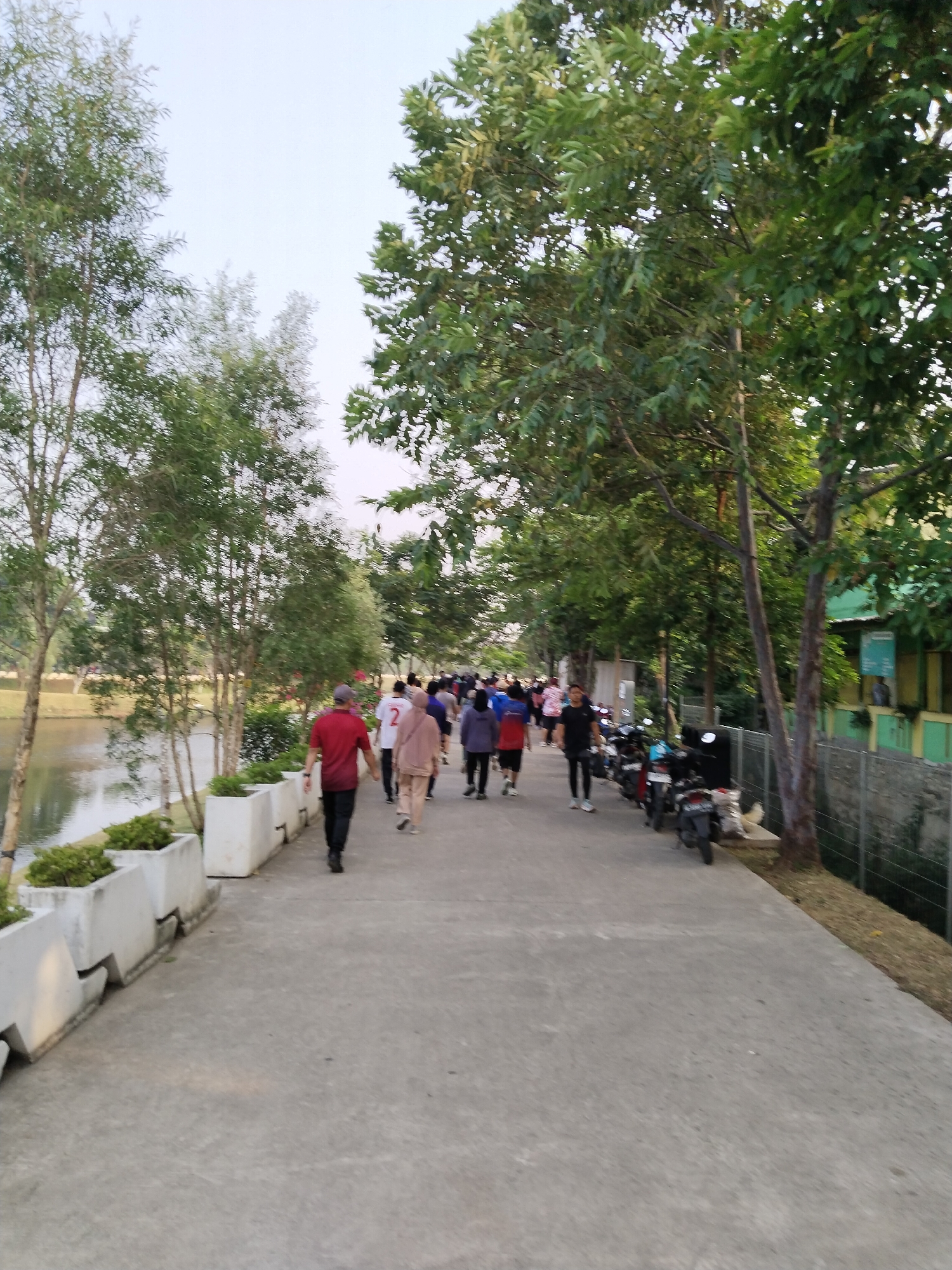 A group of people are walking and jogging on a paved path next to a body of water. The path is lined with planters and trees, and in the background, there are some buildings and parked motorcycles. The scene appears to be an outdoor recreational area in an urban setting, possibly a park or a waterfront promenade in South Jakarta, Indonesia. The lighting suggests it is either early morning or late afternoon.