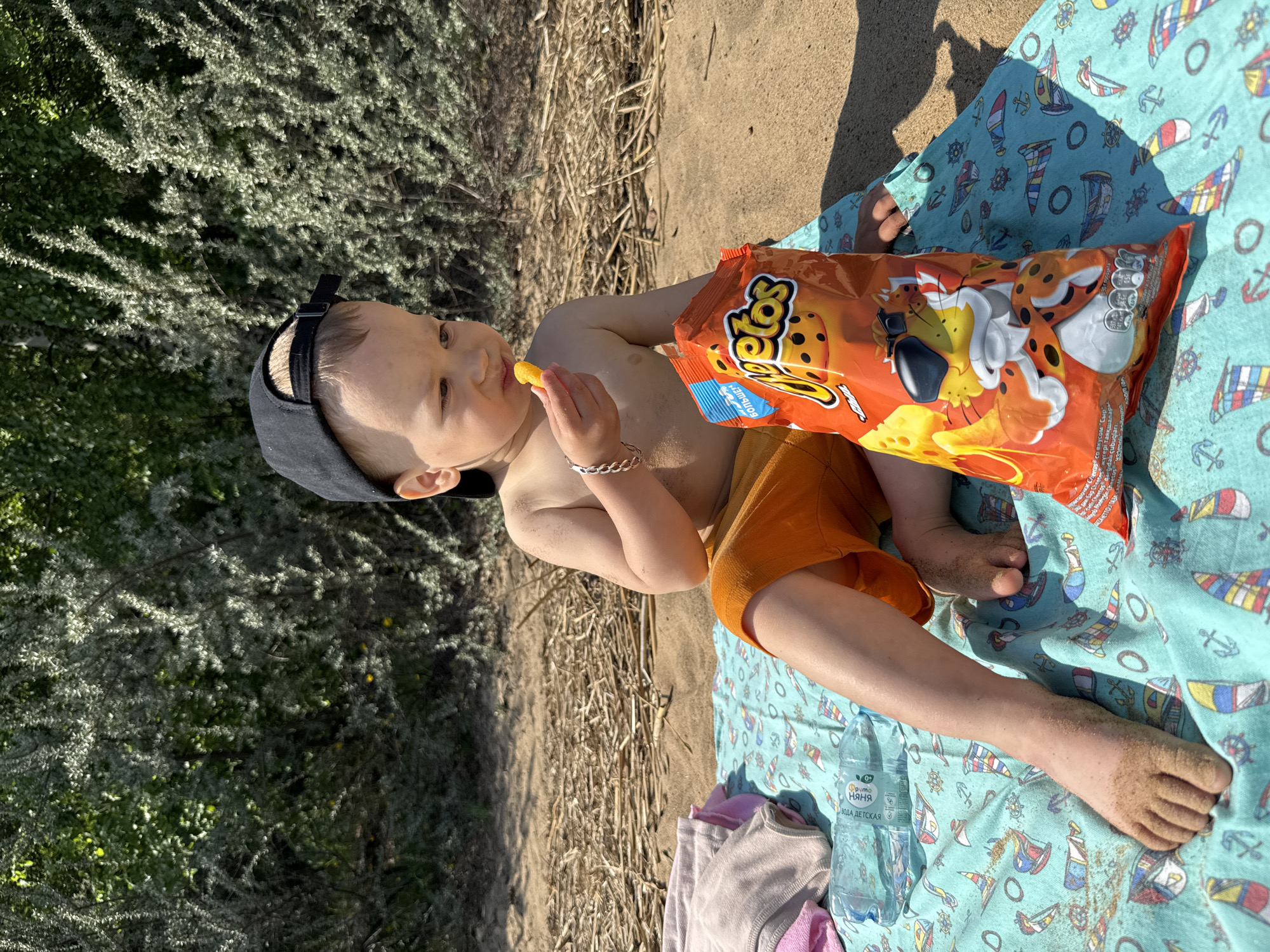 A toddler wearing a black baseball cap and orange shorts lies on a light blue blanket with nautical-themed patterns on a sandy surface. The child is holding a Cheetos snack in their hand and eating it, with a large bag of Cheetos resting beside them on the blanket. The child is wearing a silver bracelet. A bottle of water is visible near the edge of the blanket. Green foliage is present in the background. The scene appears to be outdoors, possibly at a beach or park.