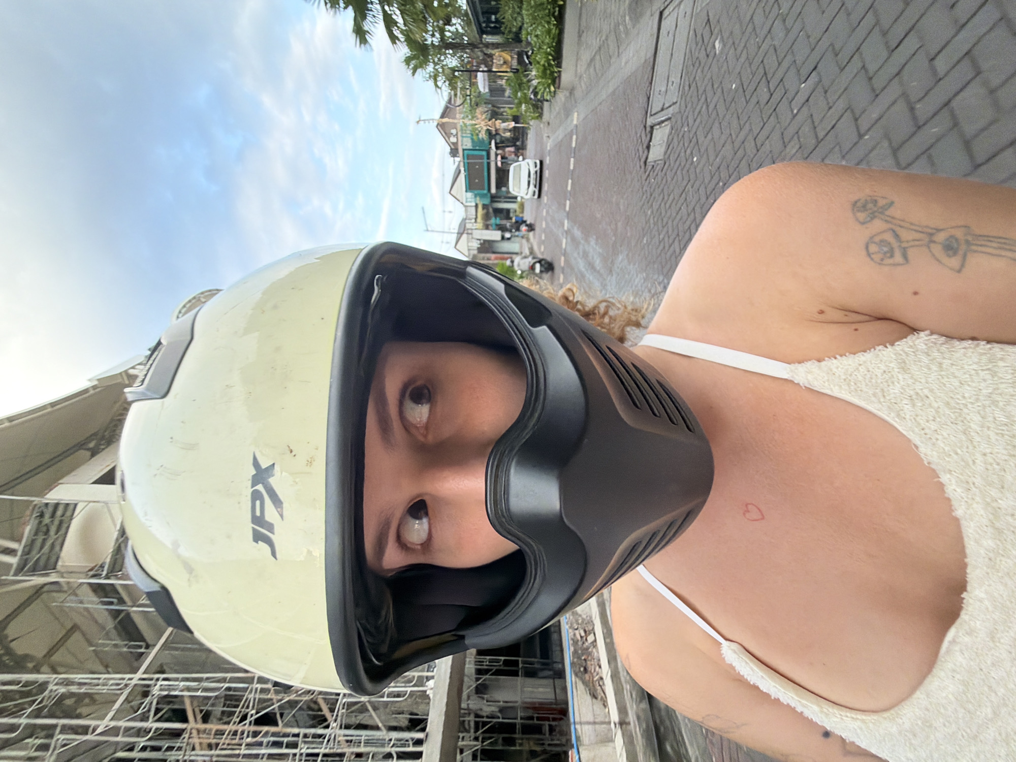 A close-up, selfie-style shot shows a person wearing a light yellow full-face motorcycle helmet with black trim and a black chin guard. The person's eyes are visible through the helmet's opening, looking to the left. They have curly blonde hair visible around the helmet. They are wearing a white tank top with a textured weave, and a small red heart tattoo is visible on their collarbone. Another tattoo of three stylized mushrooms with eyes is visible on their upper left arm. In the background, a building under construction with scaffolding is on the left. The scene appears to be a street in a tropical urban area, possibly North Kuta, Indonesia. The street is paved with gray brick. Several motorcycles and a white car are visible on the street in the middle ground. Palm trees and buildings line the street. The sky is cloudy and blue.