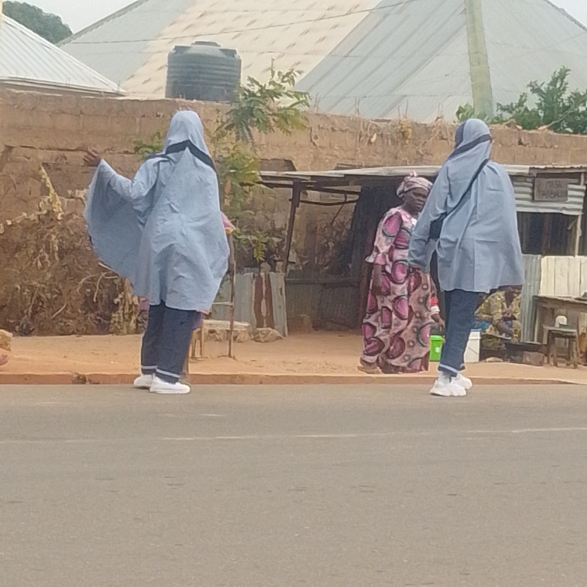 This media file depicts an outdoor scene in Gombe, Nigeria. Three individuals are visible, with two in the foreground and one in the background. The two individuals in the foreground are wearing light blue hooded garments that cover their bodies and extend down to their ankles, with dark pants and white sneakers. They are positioned facing away from the camera, with the person on the left gesturing with their left hand. The individual in the background is wearing a patterned dress and a headscarf, and appears to be standing near a structure.
The setting includes a dirt road or pathway in the foreground, with a building constructed from what appears to be mud bricks behind the individuals. Beyond the building, there are corrugated metal structures and vegetation. A water tank is visible on top of one of the buildings. The sky is overcast, suggesting cloudy weather. The lighting suggests it is daytime. A sign with some text is partially visible on a structure in the background, but it is too blurry to read. The overall impression is of a casual, everyday scene in a rural or semi-urban environment.