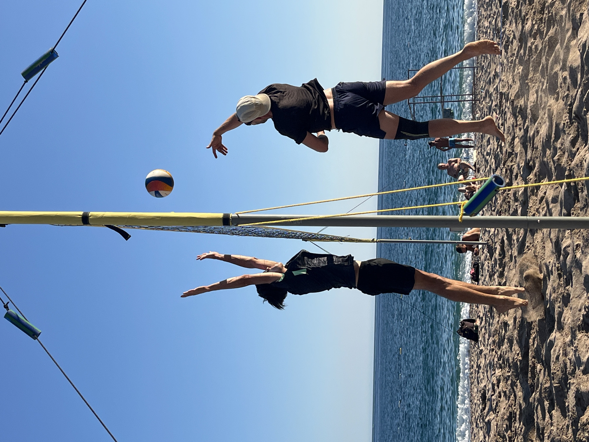 Two individuals are playing beach volleyball on a sandy beach with the ocean in the background. The person on the left is a woman wearing a black t-shirt and black shorts, jumping with her arms extended upwards. The person on the right is a man wearing a black t-shirt, dark blue shorts, and a light-colored baseball cap, also in mid-air with his arm extended to hit the ball. A volleyball is in the air between them, with the net of the volleyball court also visible. The sky is clear and blue. In the background, other people are visible relaxing on the beach and in the water. The scene appears to be a recreational activity on a sunny day. The location is identified as Zankovići, Montenegro.