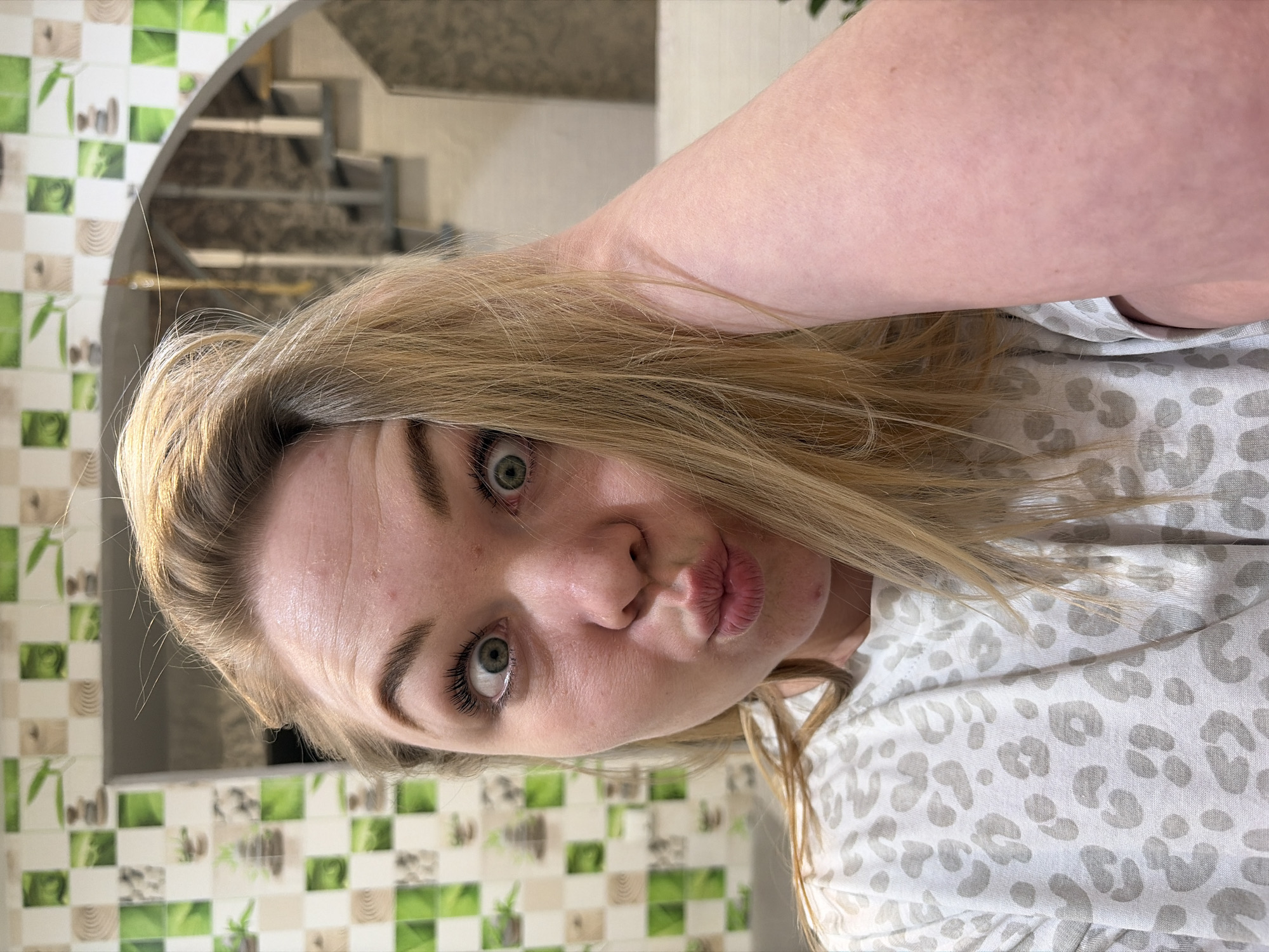 A close-up portrait features a young woman looking directly at the camera, her head tilted significantly to the left in the rotated image. She has long, straight blonde hair that falls around her face and shoulder, and wide-open light green eyes. Her lips are pursed in a playful pouting expression. She has fair skin, with visible mascara on her eyelashes and well-defined eyebrows. Minor blemishes are noticeable on her forehead and chin. She is wearing a light-colored top adorned with a subtle grey leopard print pattern, visible on her shoulder and upper torso. Her left arm, with fair skin, is bent and visible in the upper right portion of the frame.
The background is an indoor setting, likely a home or apartment in Noviy, Russia. A wall behind her is decorated with square tiles in a checkered pattern of light green, white, and a light wood-grain design; some green tiles feature a bamboo plant motif. To the right, a curved archway reveals a darker area with a staircase and metal railings in the background. The lighting suggests an indoor environment during daytime or with artificial illumination. The woman's expression conveys a sense of playfulness or mild surprise. There is no visible text in the image.