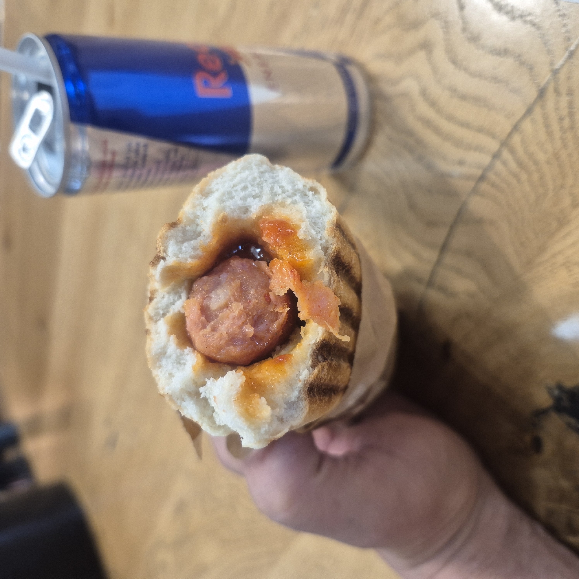 The image shows a close-up of a hot dog in a bun, held by a person's hand. The hot dog is topped with a red sauce, possibly ketchup or a spicy sauce, and some bits of what appears to be cheese or another topping. The bun is toasted with grill marks. In the background, slightly out of focus, is a blue and silver Red Bull energy drink can. The can has the prominent Red Bull logo with the bull imagery and text. The setting appears to be indoors, with a wooden surface, possibly a table, visible beneath the objects. The lighting suggests it could be daytime. The overall impression is a casual meal or snack. The text "RED BULL" is clearly visible on the can. The image was taken in Kępno, Poland.