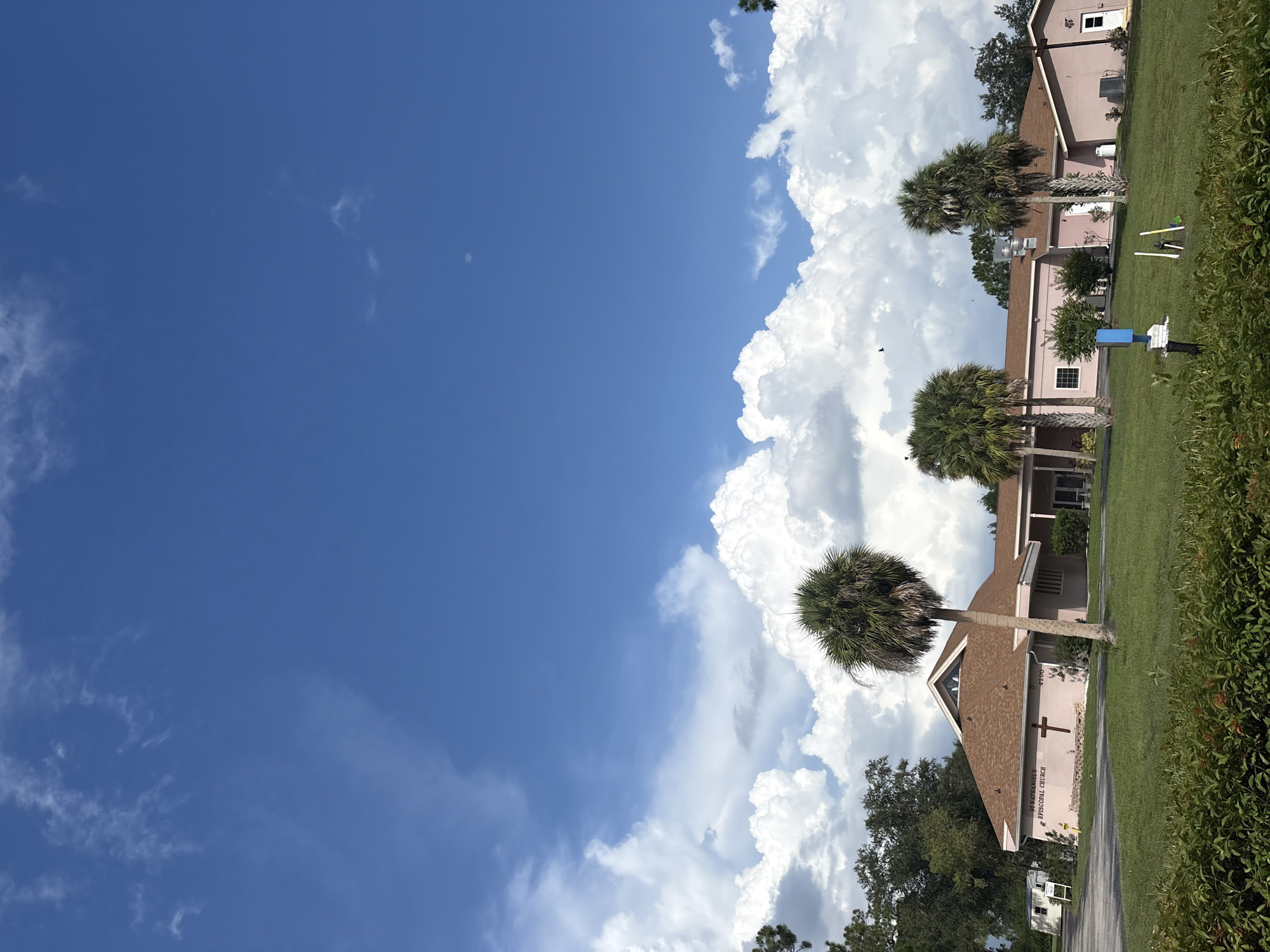 This is a full-frame, eye-level, outdoor photograph of St. Nathaniel's Episcopal Church in North Port, Florida, United States of America. The church is a single-story, pink stucco building with a brown pitched roof. A dark red cross is affixed to the front of the building. The building is situated on a well-maintained lawn with several palm trees in the foreground. The sky is a vibrant blue with large, fluffy white clouds. In the distance, a small mobile home is visible on the left side of the frame. The overall scene depicts a clear, sunny day with a few birds flying in the sky.