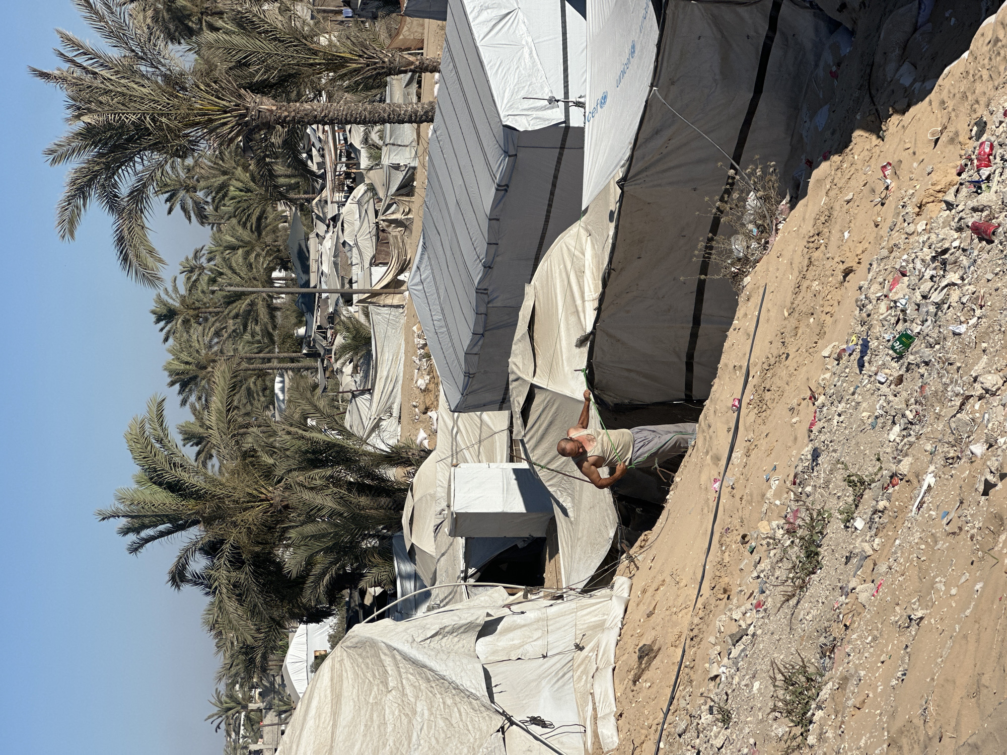 This media file captures an outdoor scene in a camp-like setting, possibly a refugee camp, given the presence of numerous temporary shelters. The shelters are constructed from white, tarp-like material and appear to be densely packed. Palm trees are a prominent feature of the landscape, with their fronds casting shadows. The ground is sandy and littered with debris, including plastic bottles and other discarded items, suggesting a somewhat unkempt environment. A man is visible in the mid-ground, leaning against a structure, with his body angled towards the viewer. He is wearing a light-colored tank top and grey pants. The lighting suggests it is daytime, with the sun casting distinct shadows. The sky is clear and blue. One of the shelters has "UNICEF" written in blue text on its side. The overall impression is one of a functional, yet potentially challenging, living situation.