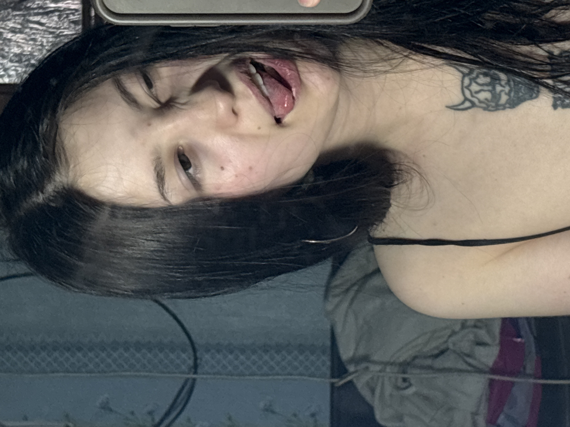 A woman with dark hair and a shoulder tattoo is visible in a close-up shot. Her mouth is open, and her head is tilted to the side. She is wearing a thin black strap, possibly from a garment. The background appears to be a patterned surface. There is no contextual information available to determine if the photo was taken in Фоки, Russia or to identify the environment with any accuracy.