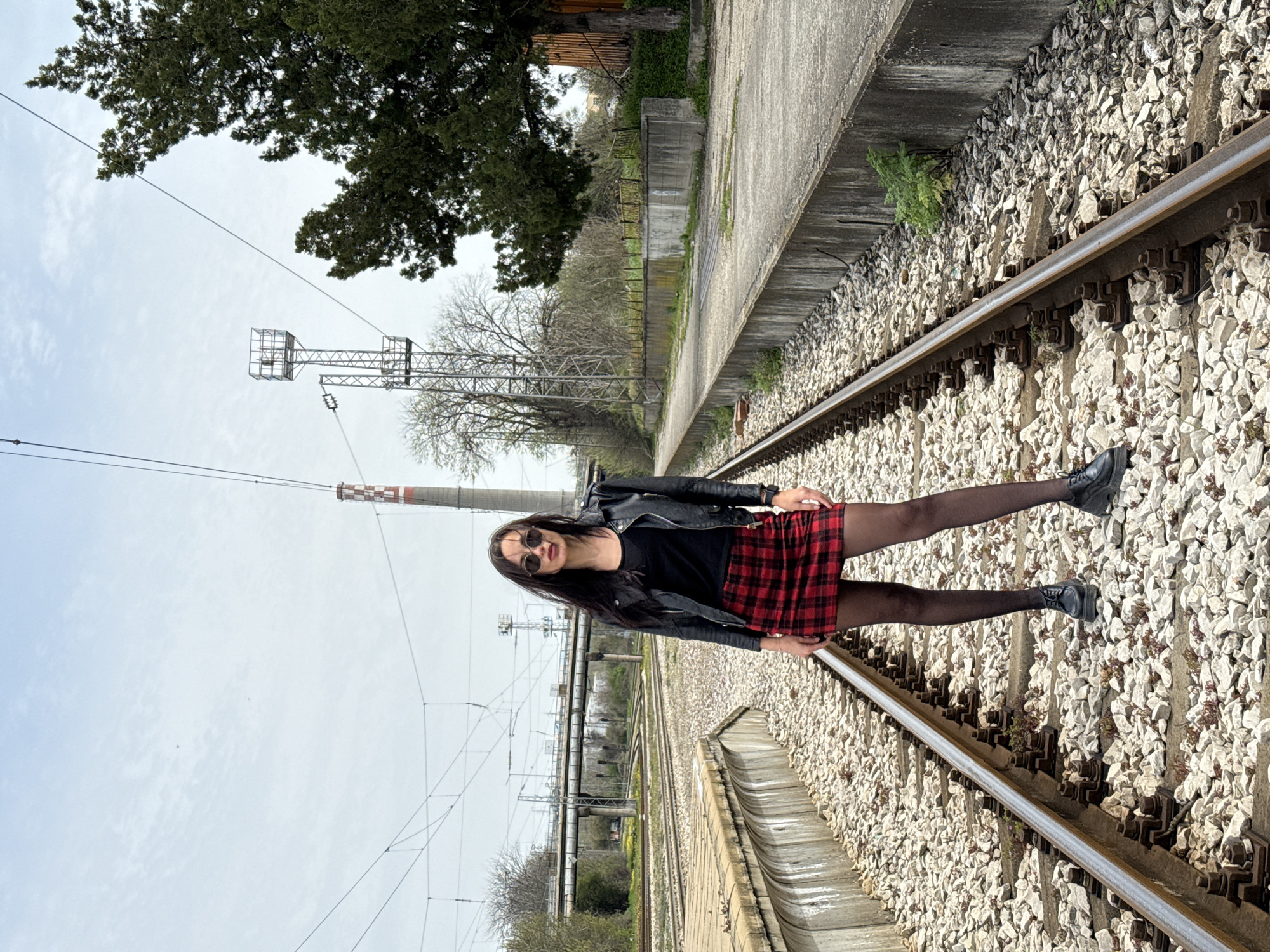 A young woman with long dark hair stands on a railway track in Pleven, Bulgaria. She is wearing a black leather jacket over a black top, a red and black plaid skirt, black stockings, and black boots. The railway track is lined with light gray gravel. Overhead, there are utility poles and power lines. Trees and vegetation are visible along the side of the railway. The sky is overcast.