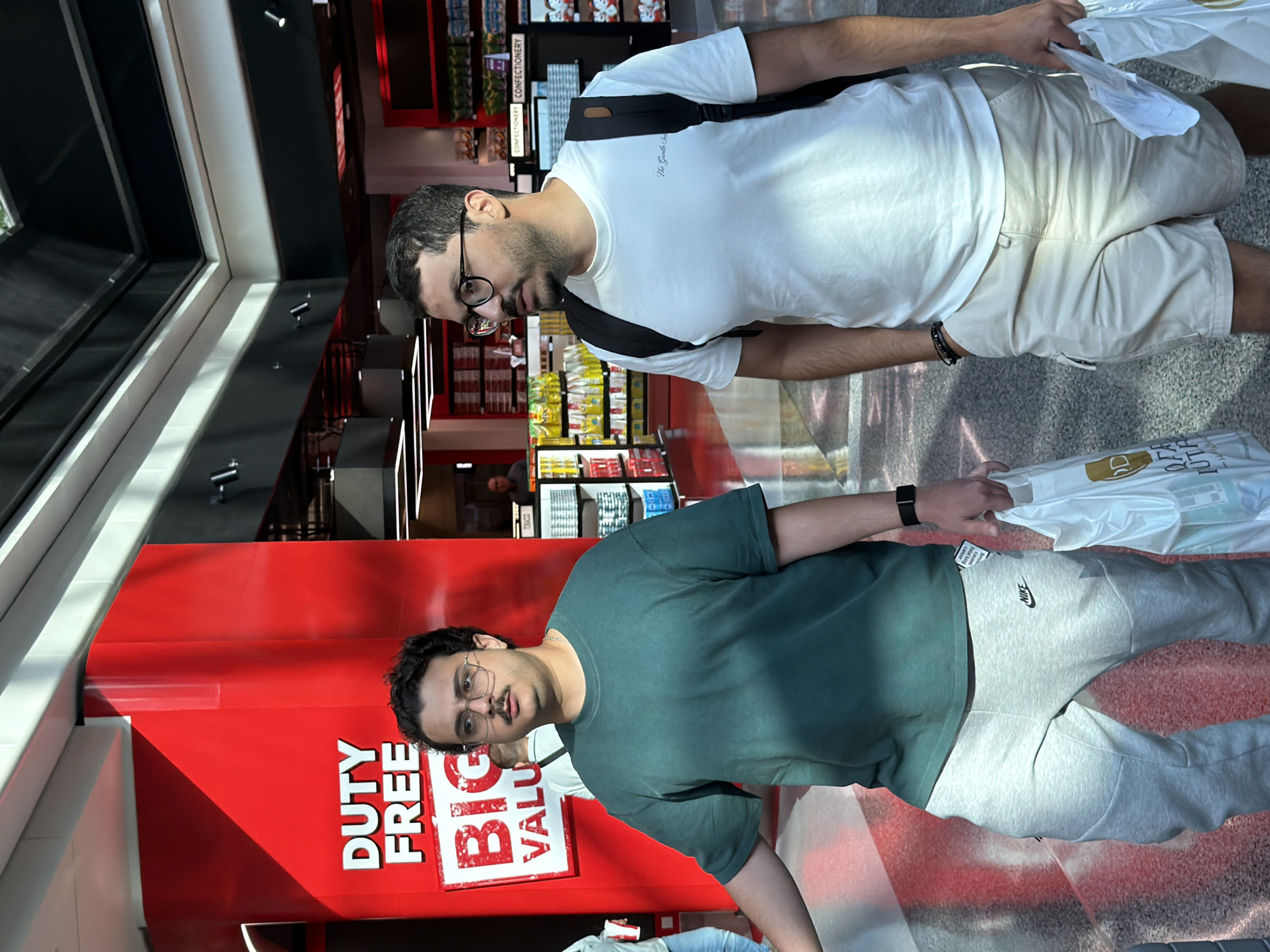 Two men are walking in an airport terminal. The man on the left is wearing a green t-shirt and gray sweatpants. He has glasses and a mustache. The man on the right is wearing a white t-shirt and beige shorts. He has glasses and a beard and is carrying a black backpack. Both men are holding shopping bags. In the background, there is a large red sign that says "DUTY FREE BIG VALUE." There are also shelves stocked with products, suggesting a retail area. The lighting and architecture indicate an indoor public space, consistent with an airport. The presence of "DUTY FREE" signage points to a location within an international airport.