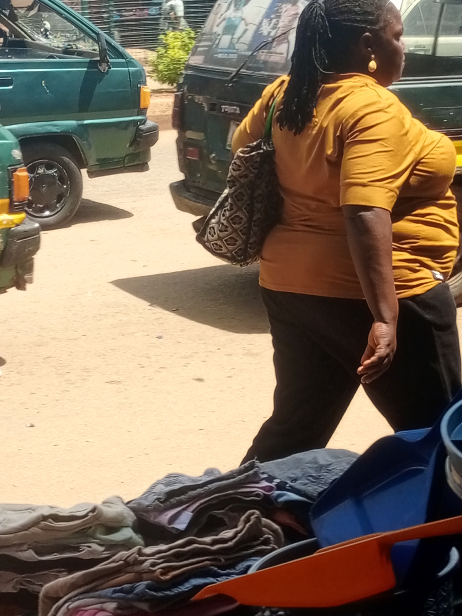 In Bukuru, Nigeria, a woman in an ochre t-shirt and black trousers walks past a green van. She carries a patterned bag over her shoulder and wears a gold earring. In the foreground, a pile of folded clothing sits next to blue and orange plastic containers. The ground is sandy, and the scene is illuminated by sunlight, casting shadows.