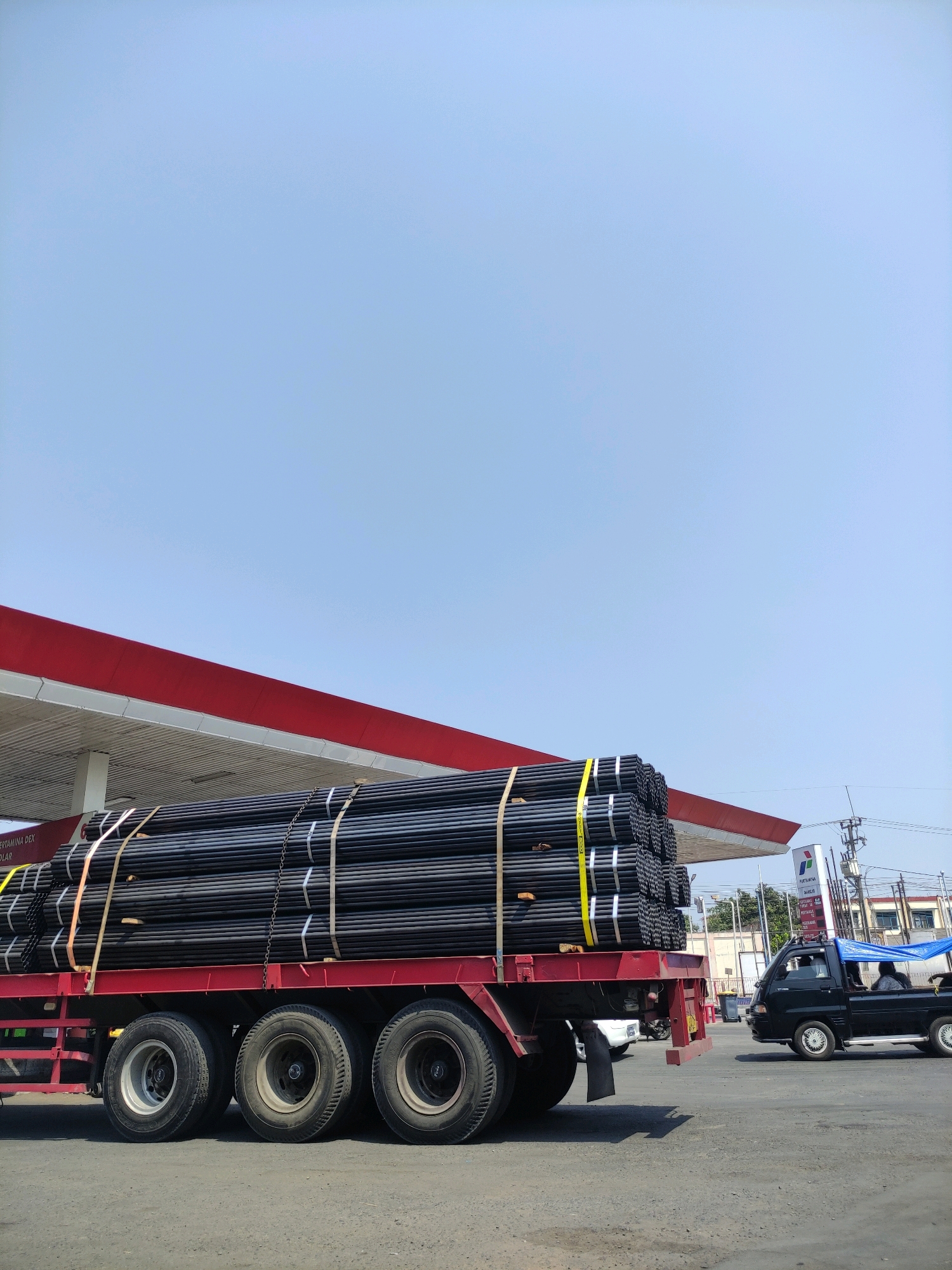 A large truck is parked at a gas station in Cadangpinggan, Indonesia. The truck is loaded with numerous black pipes, bundled together with yellow and white straps. The truck has a red trailer and multiple tires. In the background, a red and white awning of the gas station is visible, along with a Pertamina sign. A black pickup truck with a blue tarp is driving away from the gas station, and two people are seated inside it. The sky is clear and blue.