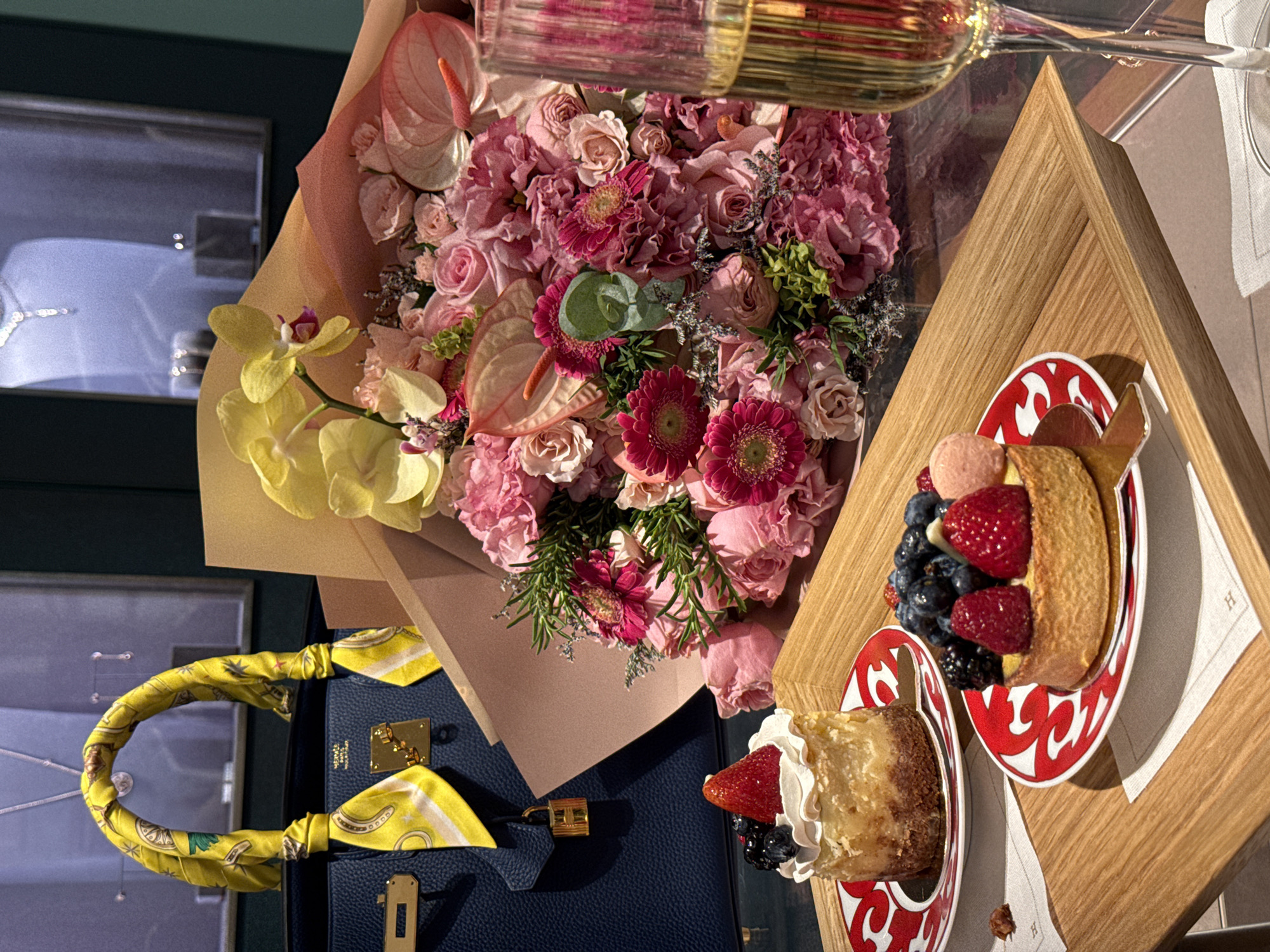 A still life image shows a blue leather handbag with a yellow scarf tied to its handle, a bouquet of pink and yellow flowers, two desserts on red and white patterned plates, a champagne flute, and a wooden tray. The handbag and flowers sit near a window display of jewelry. The desserts are topped with strawberries, blueberries, and whipped cream. The image was taken in the city of Zapopan, Mexico.