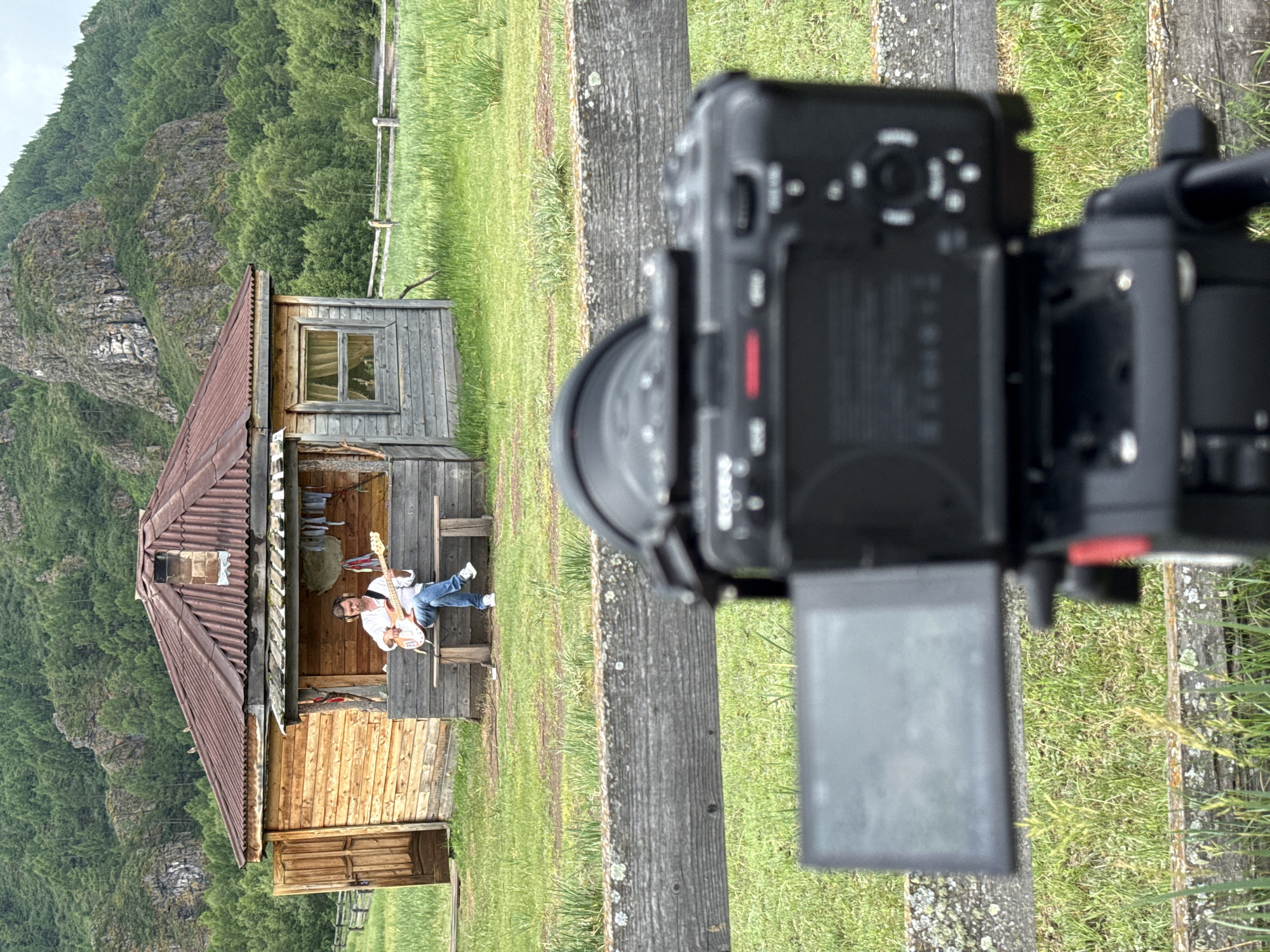 In Anchyl-Chon, Russia, a man is playing a guitar while sitting on a wooden bench in front of a rustic wooden house. The house has a red metal roof and a porch. The man is wearing a white shirt and blue jeans. He is also wearing headphones. The background shows a grassy field and a forested mountain. In the foreground, a camera on a tripod is pointed at the man, suggesting the scene is being filmed.