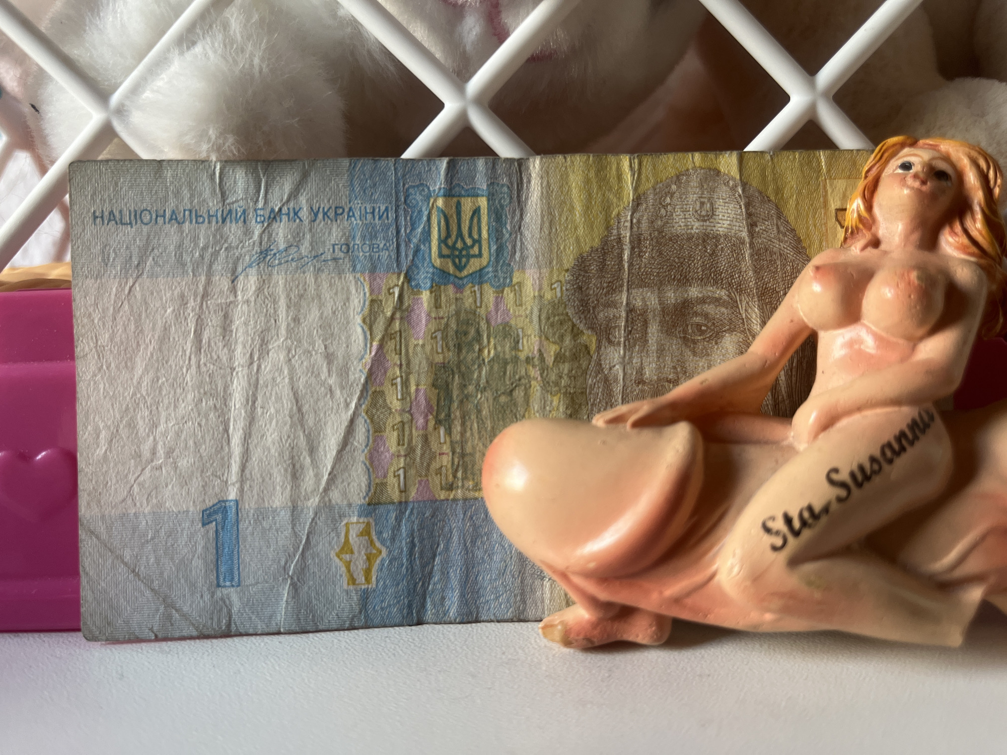 The image shows a close-up of a Ukrainian 1 Hryvnia banknote. A small, pink-toned figurine of a nude woman is positioned on the right side of the banknote, partially obscuring it. The figurine has blonde hair and is depicted in a reclining pose. The word "Sta. Susanna" is written in black cursive on the figurine's leg.
The banknote itself displays Ukrainian national symbols, including the trident emblem and text in Ukrainian. The number "1" is prominently visible on the left side of the banknote. The background includes a white, lattice-like structure and some soft, fluffy material, possibly a blanket or stuffed toy, suggesting an indoor setting. A bright pink object with a heart shape is also partially visible on the far left. The lighting appears to be artificial, casting soft shadows. The scene has a playful and somewhat provocative tone due to the juxtaposition of the currency and the figurine. No specific time of day or weather conditions can be determined. The location context of Schwäbisch Hall, Germany, is not visually indicated within the image itself.