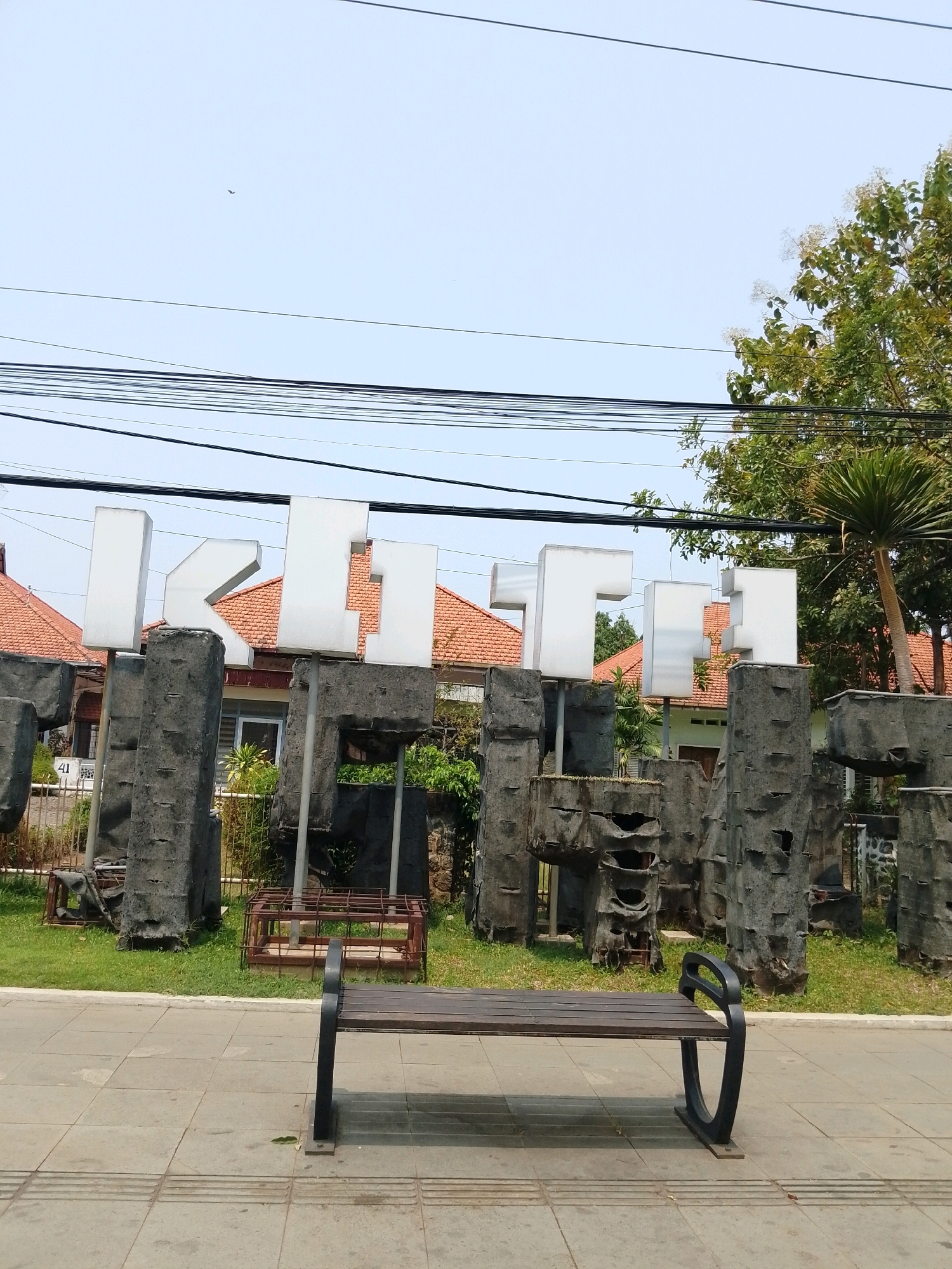 The image shows a large, white, three-dimensional sign spelling "IKITII" in Pasuruan, Indonesia. The letters are mounted on rough, dark gray concrete pillars that resemble natural rock formations, some with open, hollowed-out sections. Behind the sign, houses with red tile roofs are visible, as well as lush green trees and a palm tree. The foreground features a paved walkway with a wooden bench in front of it. A patch of green grass is between the walkway and the sign. Overhead, power lines crisscross the sky, which is a clear, pale blue. The overall scene suggests a public space or entrance area.