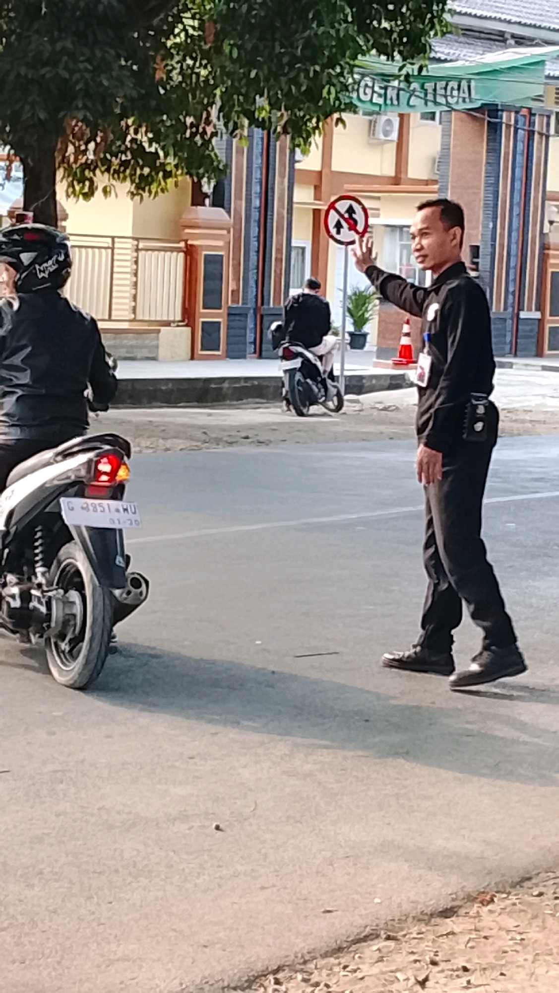 In the city of Slawi, Indonesia, on a street, a man in a black uniform is directing traffic. He is standing in the road, facing left, with his right arm raised and his index finger extended. He appears to be a traffic officer or security guard. To his left, a motorcycle with a rider wearing a black helmet and jacket is in the foreground. In the background, another person on a motorcycle is further down the street, and a building with a sign that reads "GER 2 TEGAL" is visible. A traffic sign indicating "no U-turn" is also present. The lighting suggests it is daytime, and the weather appears to be clear.