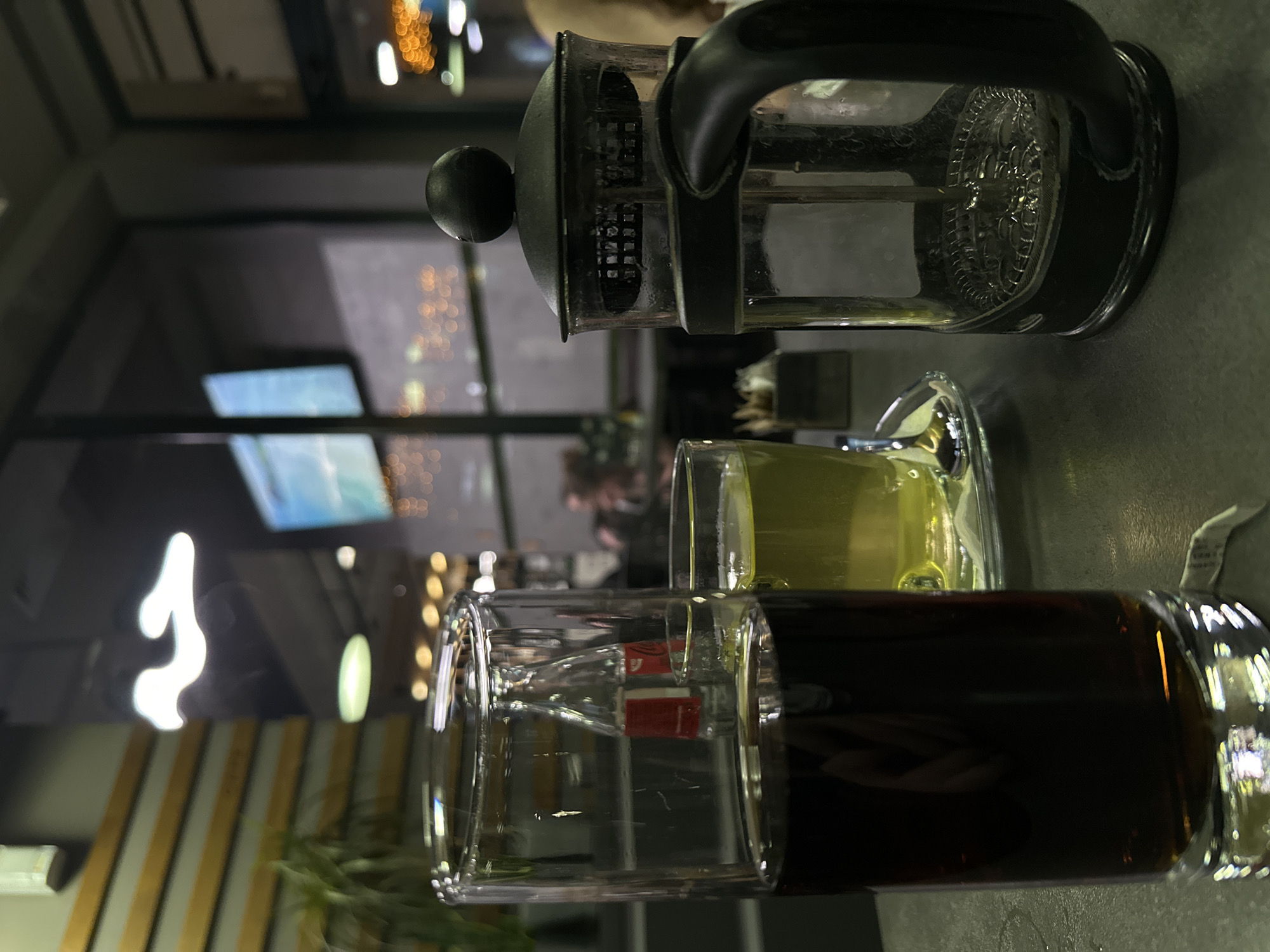 The image shows a French press with a black handle and a clear glass body, positioned on a table in what appears to be a café or restaurant. Next to the French press, there are two glass cups; one contains a light-colored liquid, possibly tea, and the other contains a dark liquid, which could be coffee or another type of tea. The table surface is gray. In the background, there's a blurred interior view of the establishment, including lighting, wall decor with white and yellow stripes, and what seems to be a television screen. The presence of the French press, along with the two beverages, suggests that this scene is located in a café or a restaurant setting where hot beverages are being served. While I can discern that the location is likely a café, based on the available visual clues, I cannot confirm whether this is specifically in Stara Zagora, Bulgaria.