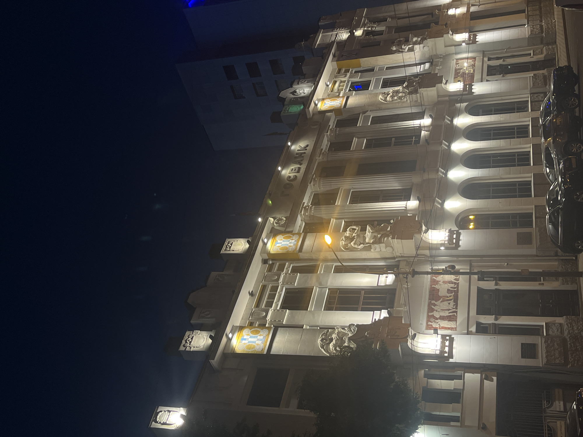 This is a nighttime photograph of the façade of the GOSBANK building in Krasnodar, Russia. The building is illuminated by exterior lights, highlighting its ornate architectural details. The image is oriented vertically, with the night sky dominating the left side and the building filling the right.
The building features multiple stories with large arched windows on the lower floors and rectangular windows on the upper floors. Decorative elements, including sculptures and decorative panels, adorn the façade. One panel displays a mosaic-like design with yellow and blue squares, while another features a frieze-like depiction of figures in white against a reddish background.
Several streetlights are visible, casting pools of light on the building and the surrounding area. A few cars are parked along the street in front of the building, their shapes partially visible due to the lighting. A tree with dark foliage is visible in the lower right portion of the image, obscuring some of the building's base.
The overall mood is serene and slightly mysterious, attributed to the nighttime setting and the dramatic illumination of the historic architecture. The visible text on the building, "ГОСБАНК," is the Russian abbreviation for State Bank. The time of day is clearly night, and the weather appears to be clear, as there are no indications of rain or fog.